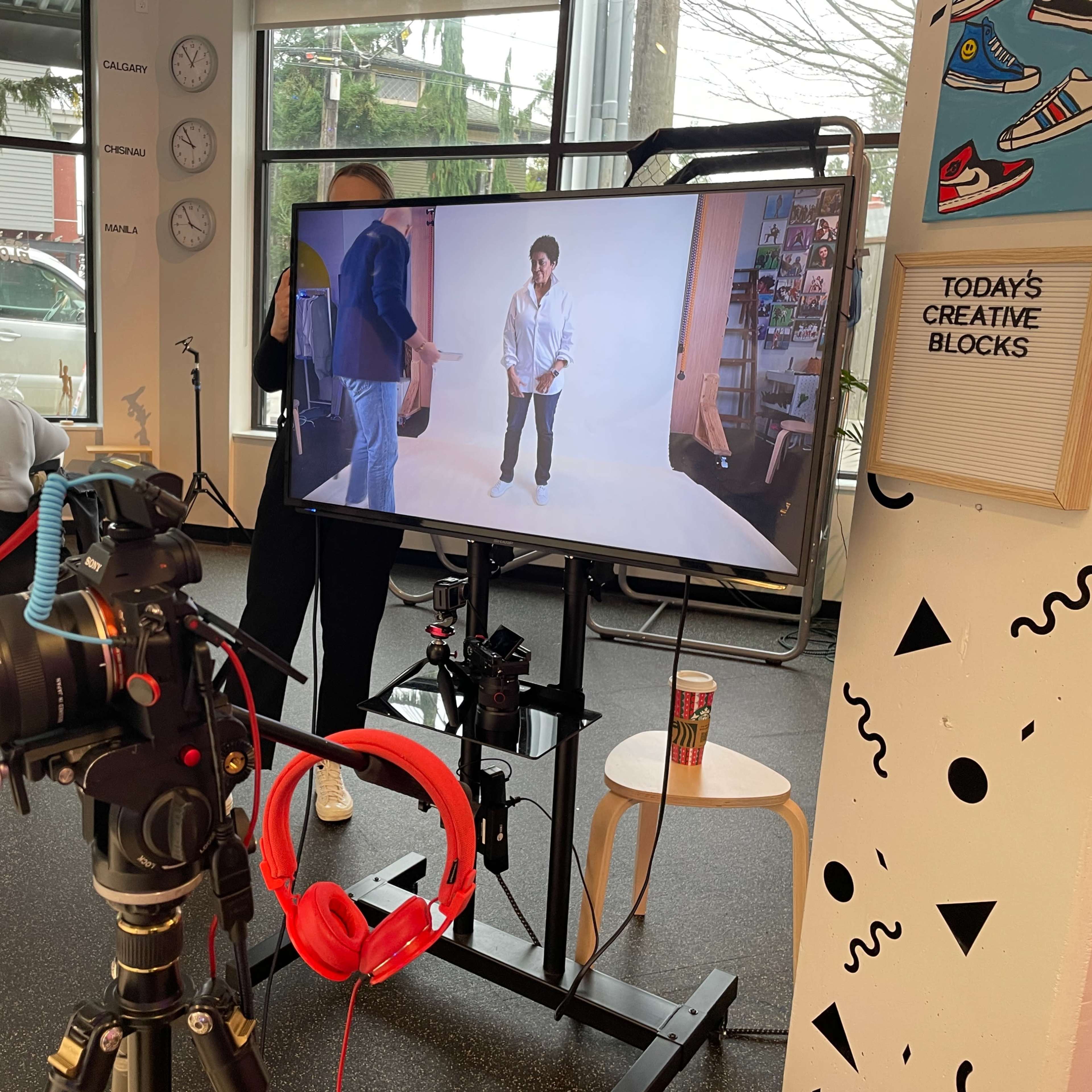 A camera on a tripod is positioned in front of a television screen displaying a person posing, while people work at tables in the background.