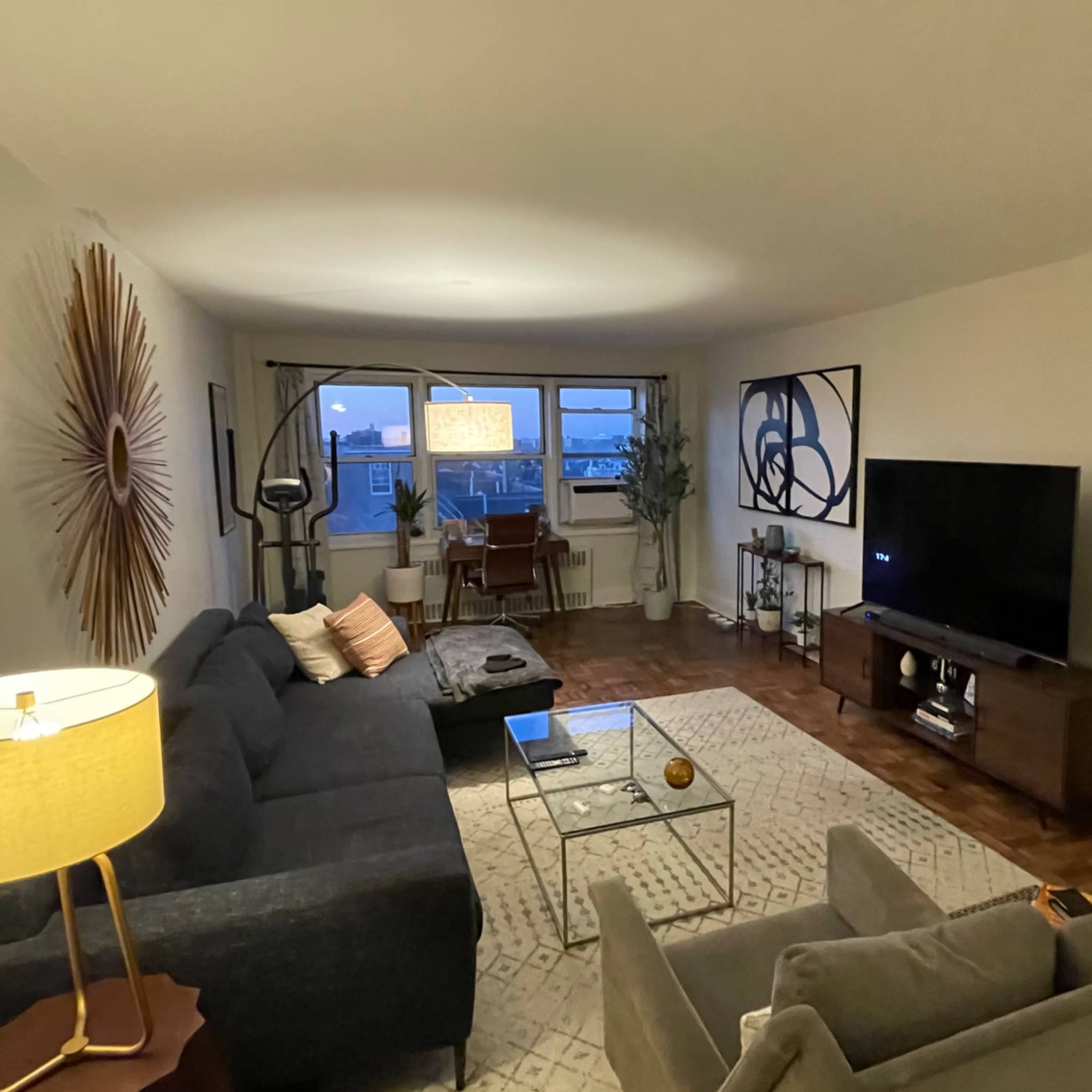 The living room features a large gray sectional sofa, a glass coffee table, a flat-screen TV on a wooden stand, and a window with a view of the outdoors.