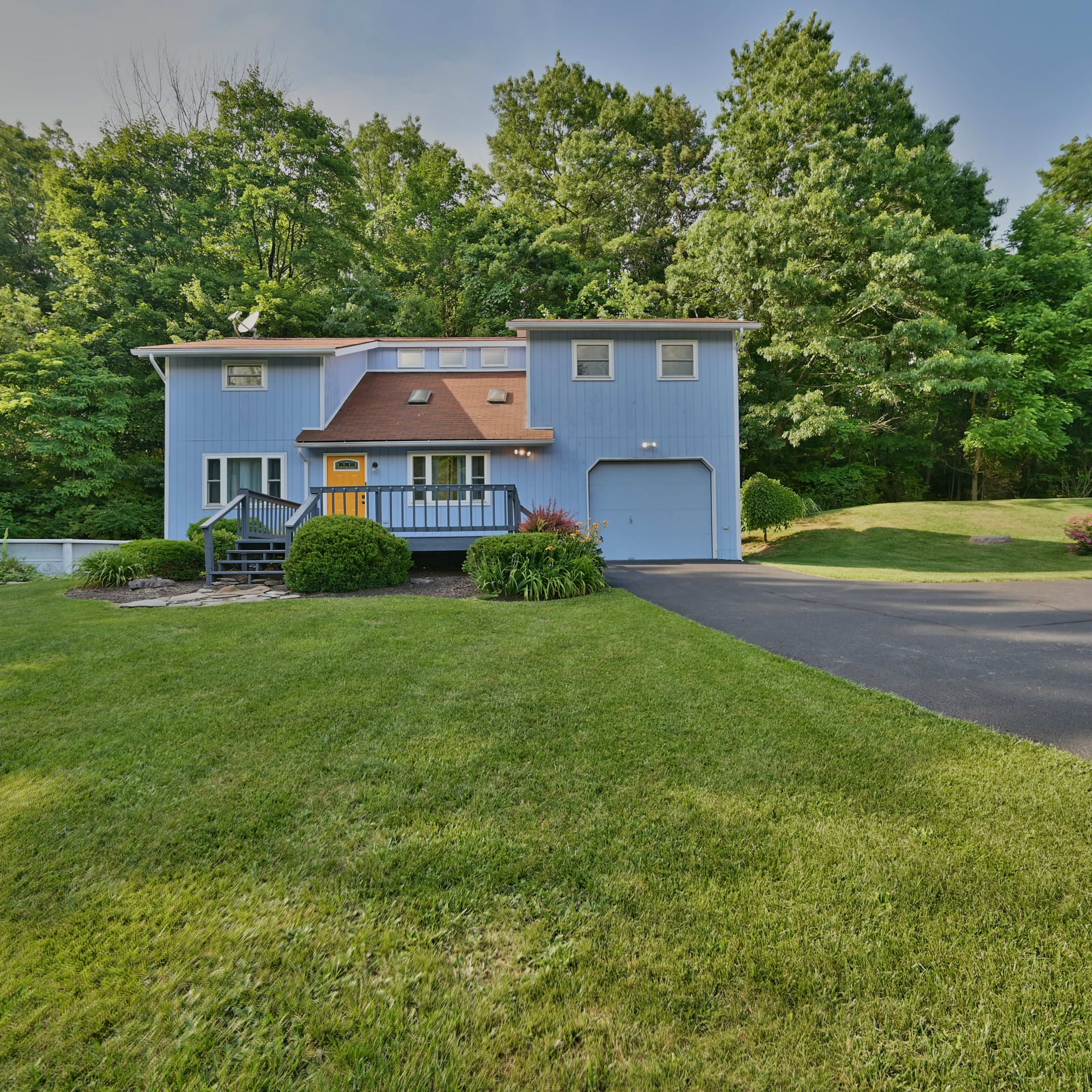 A two-story blue house with a brown roof sits on a green lawn, surrounded by trees and a paved driveway.