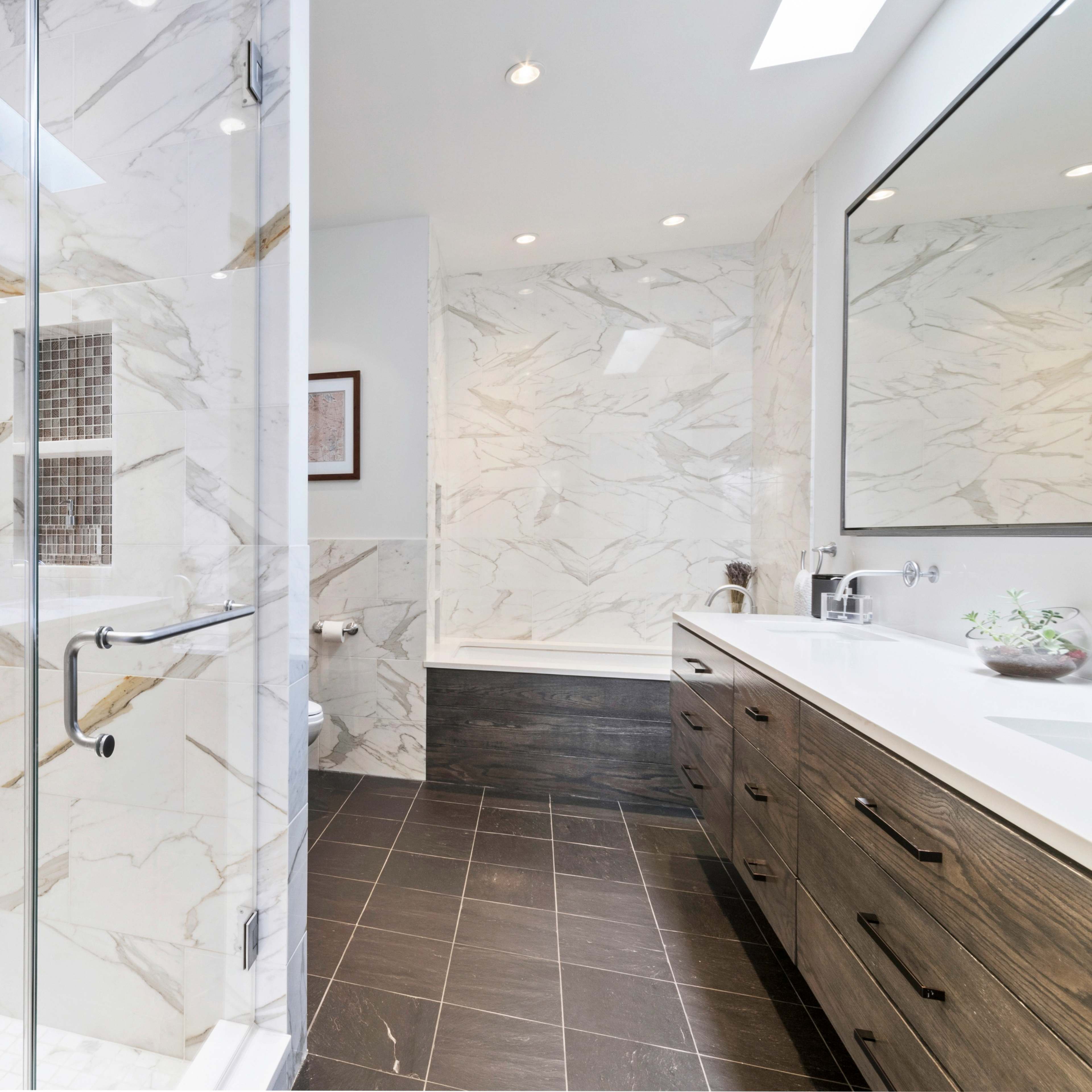 A modern bathroom featuring a glass shower, marble walls, and a double vanity with wooden cabinetry.