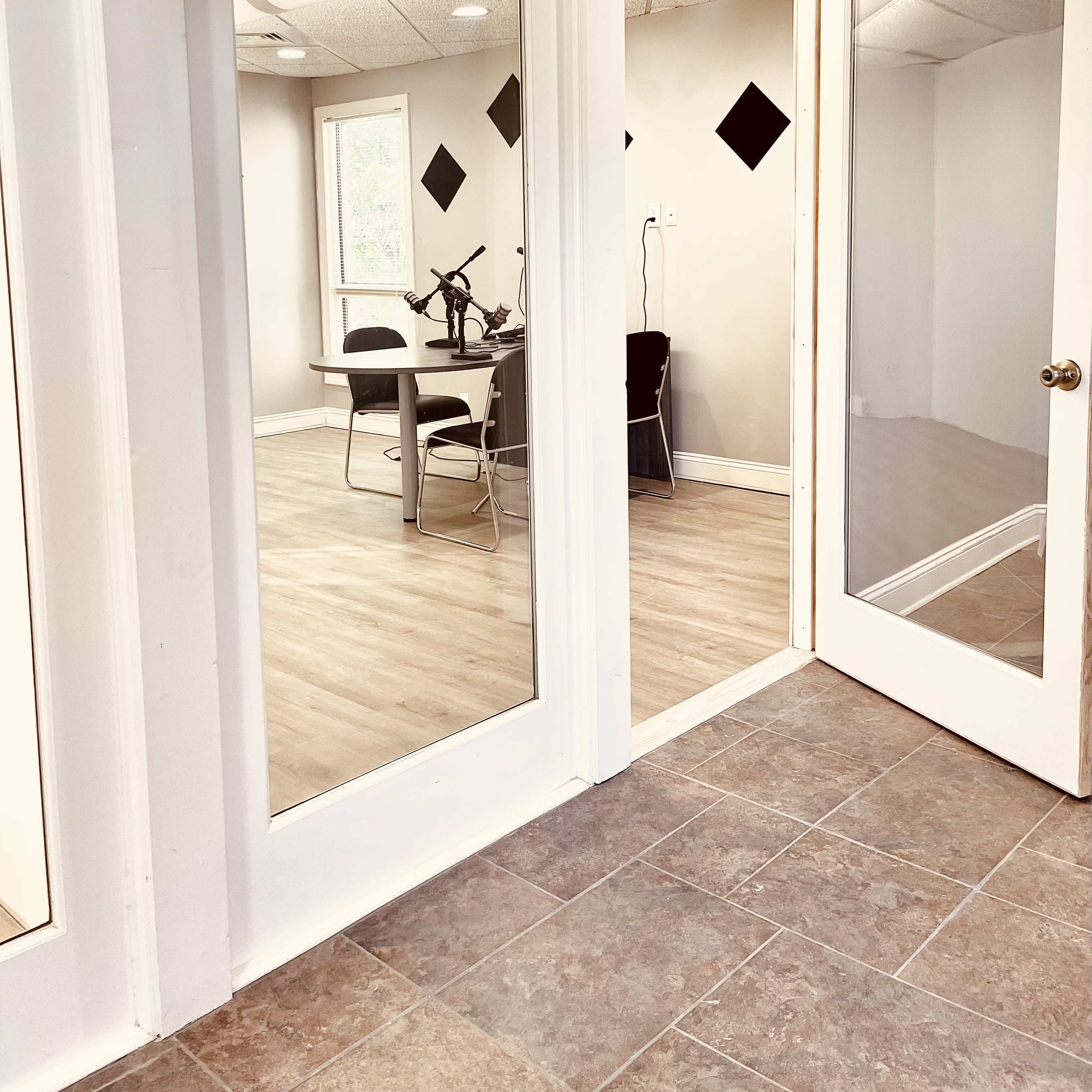 A corridor with two large glass-paneled doors leading into a conference room, featuring a table and chairs, against a backdrop of light-colored walls and a wooden floor.