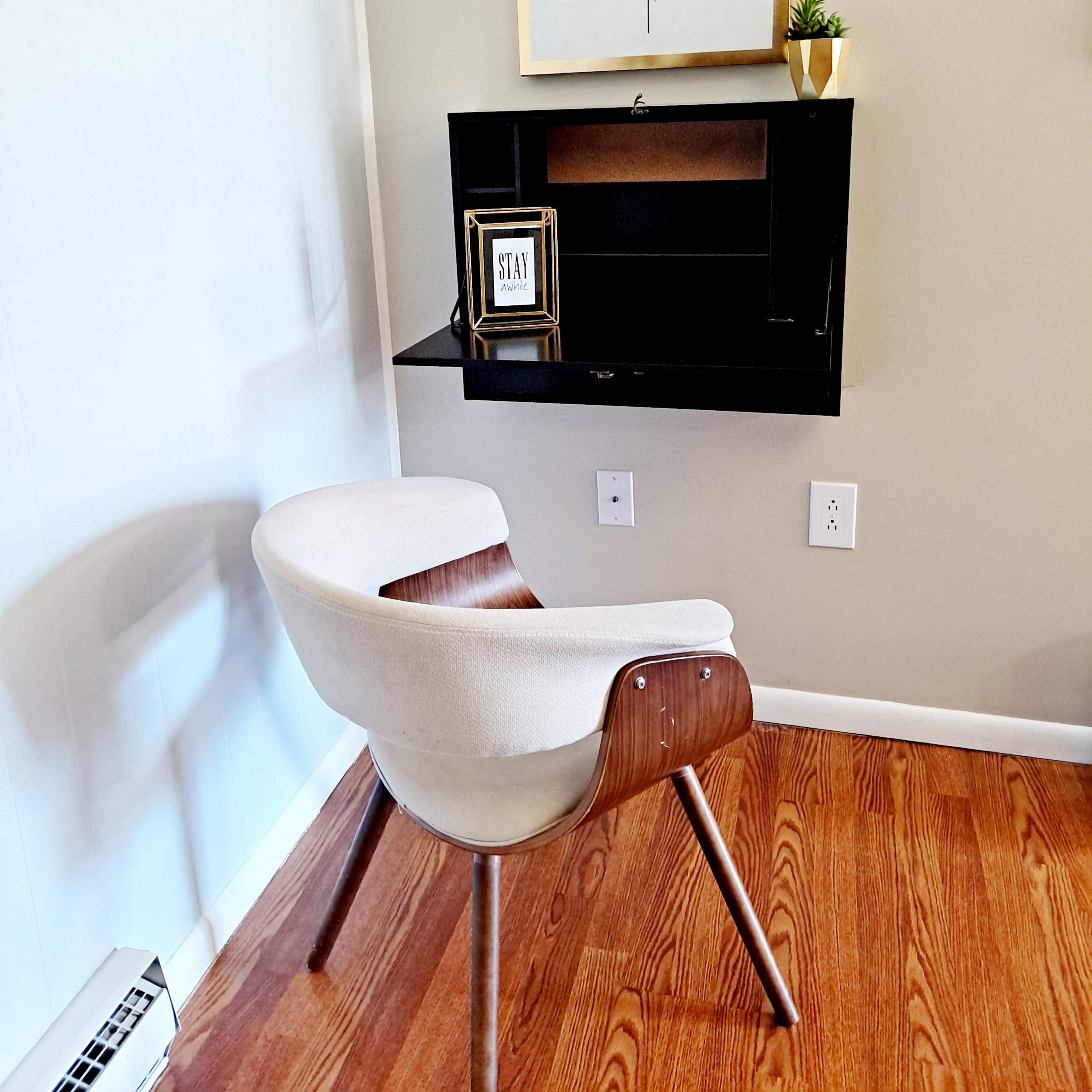 A wooden chair is positioned next to a wall-mounted desk, with a framed motivational quote and a small plant above it.