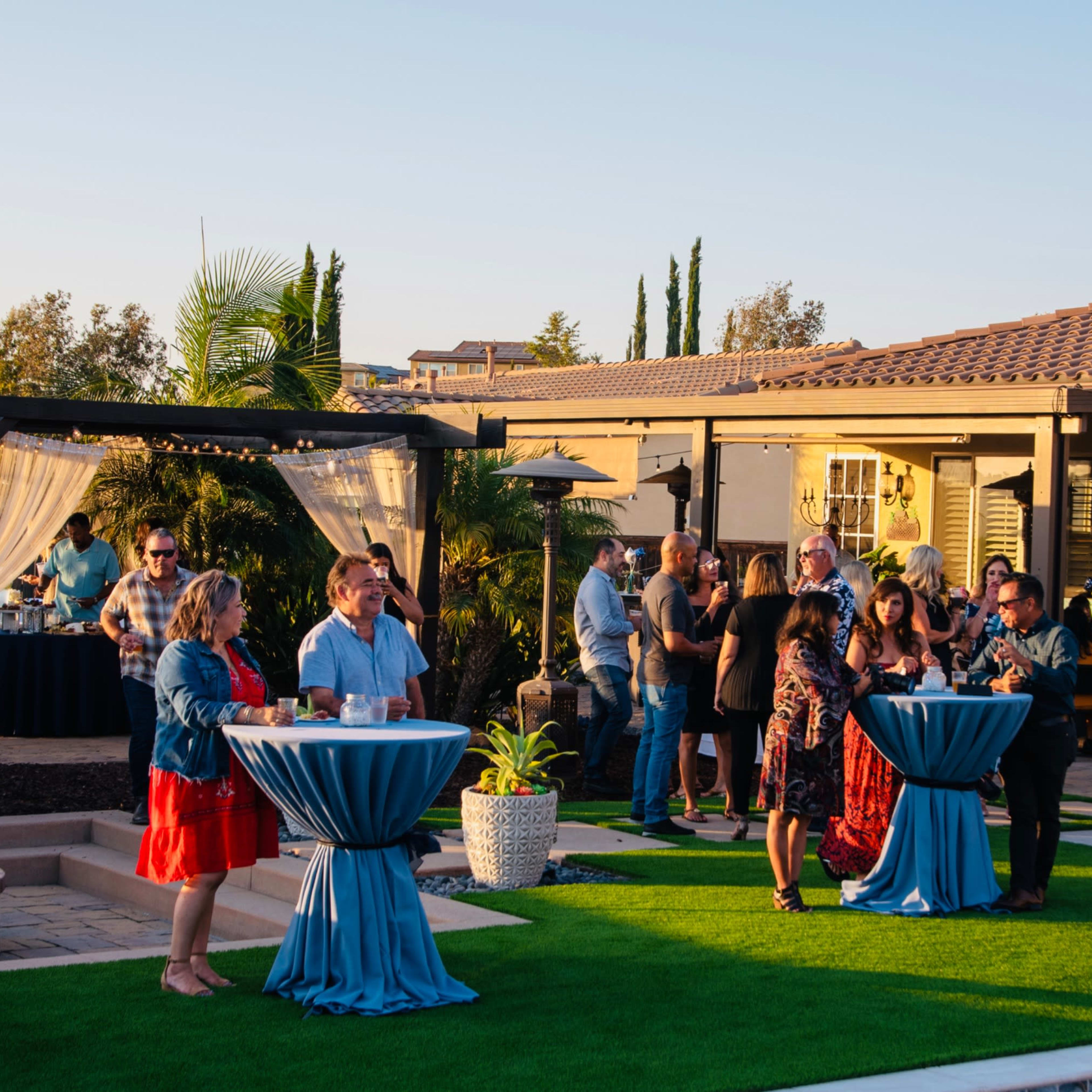 A group of people socializes around blue-draped tables in a landscaped outdoor setting during a gathering.