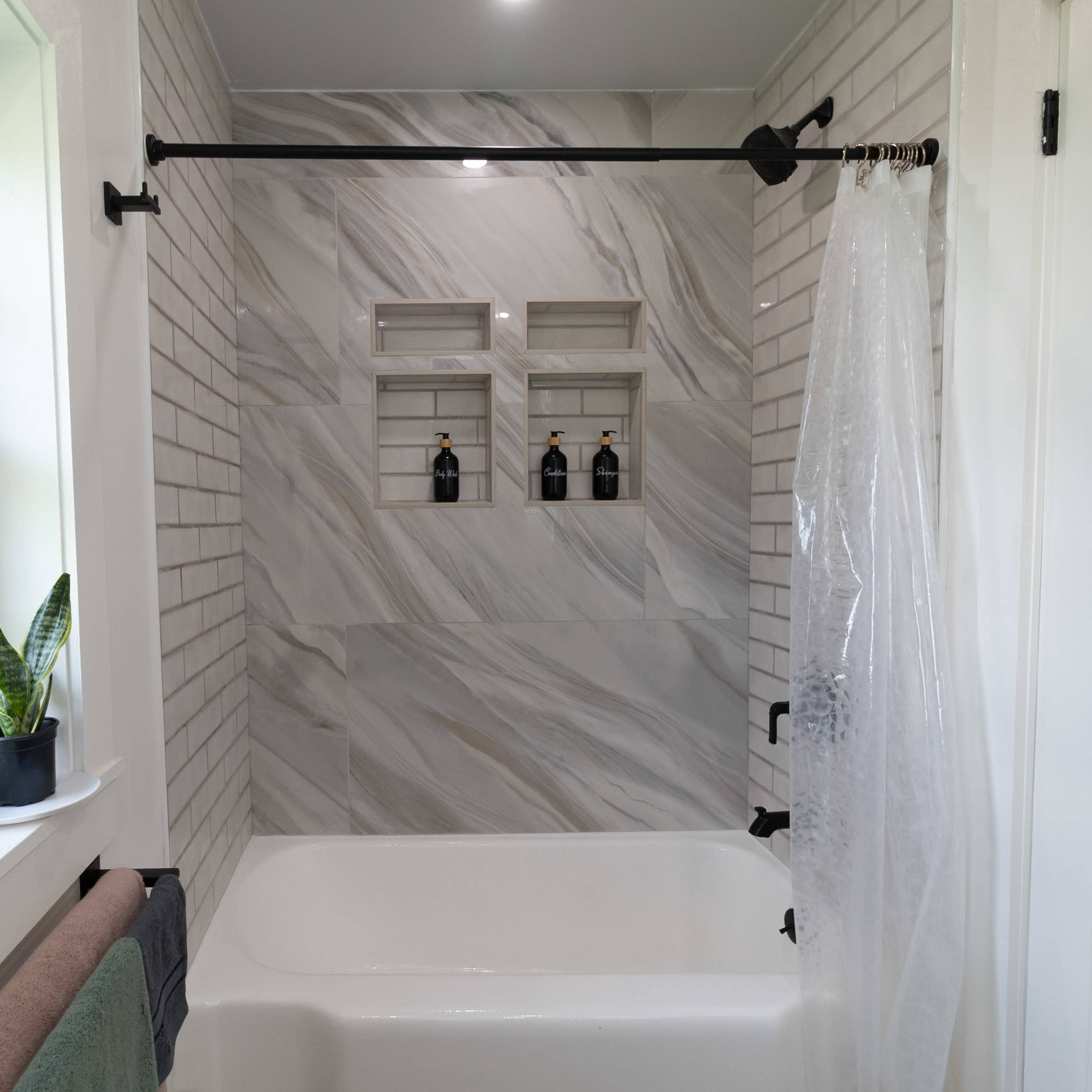 A bathroom featuring a bathtub against a wall with gray marble tiles and built-in shelves displaying three decorative bottles.