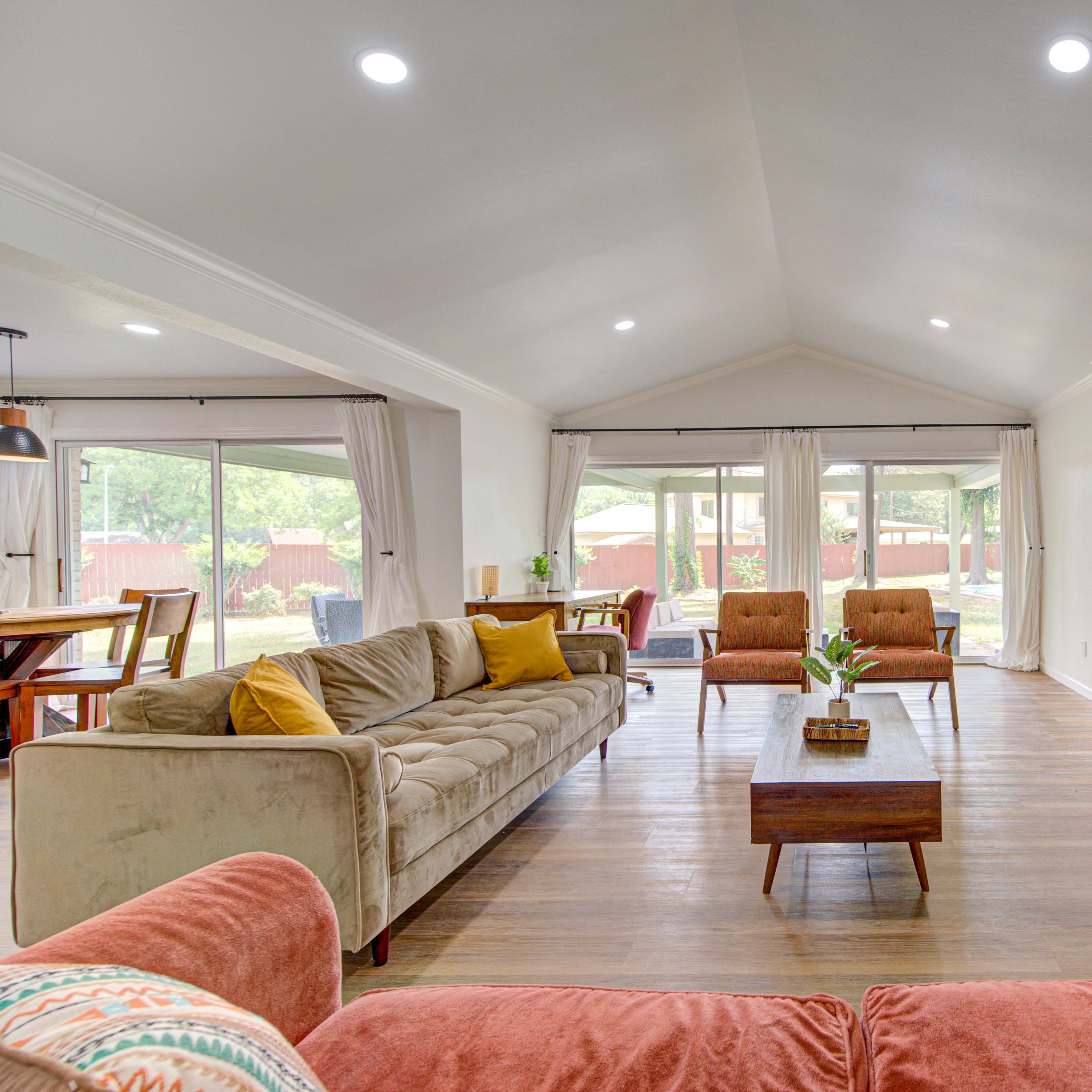 A spacious living room with a light-colored sofa, two armchairs, a wooden coffee table, and a television, set against large windows that provide a view of an outdoor patio.
