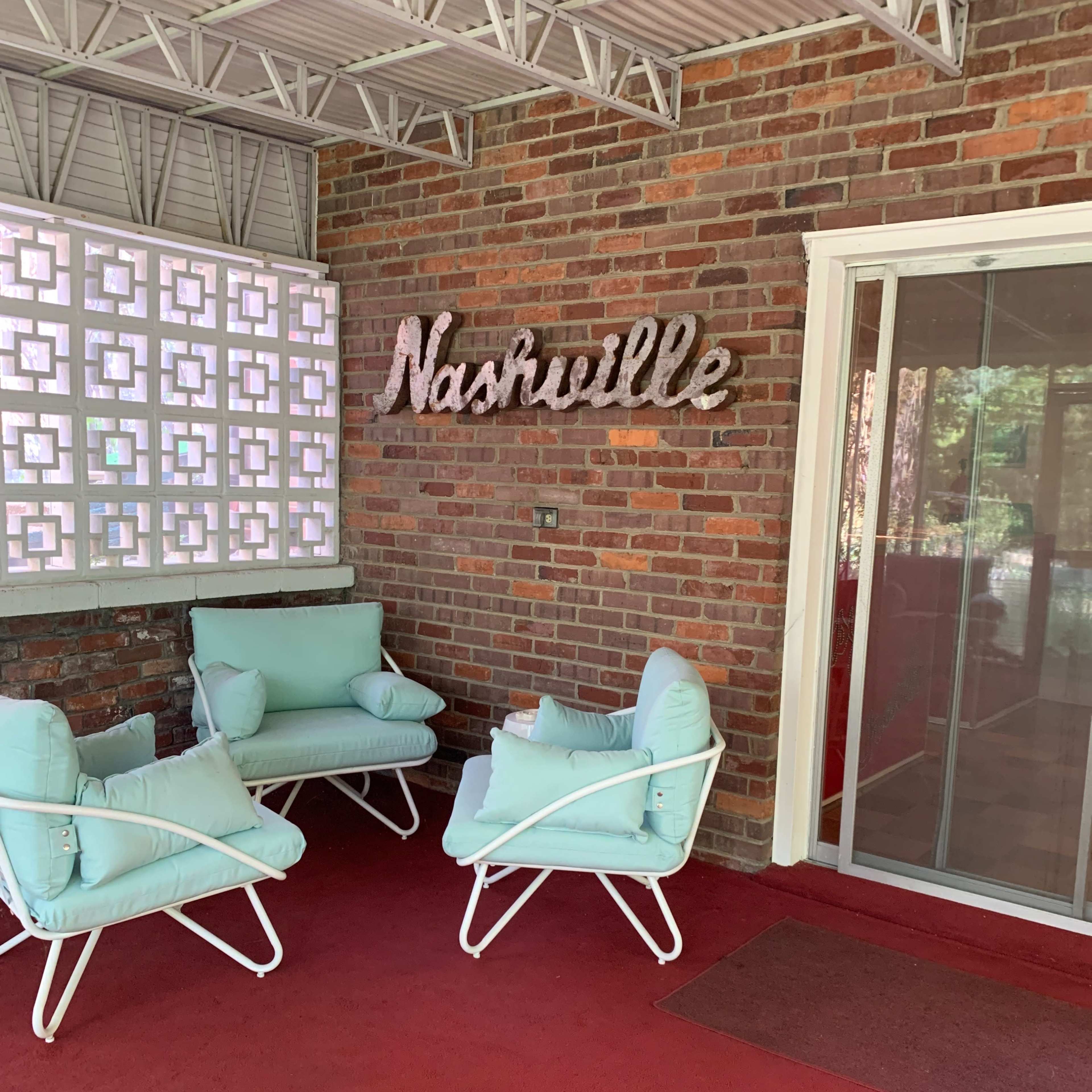 A covered patio features three light blue chairs arranged around a red carpet, with "Nashville" sign displayed on a brick wall.