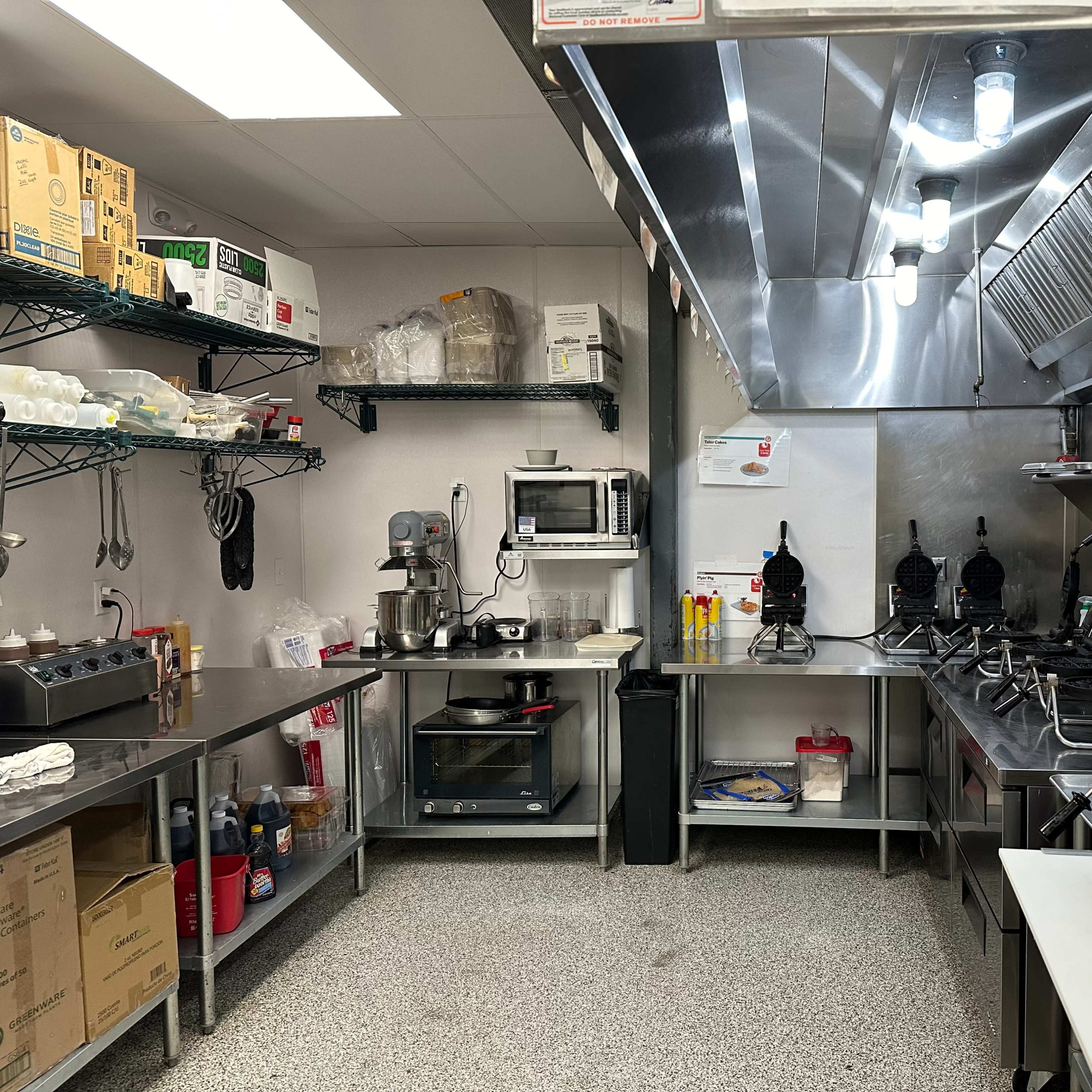 A commercial kitchen featuring stainless steel countertops, an oven, a microwave, and multiple waffle makers on the right side.