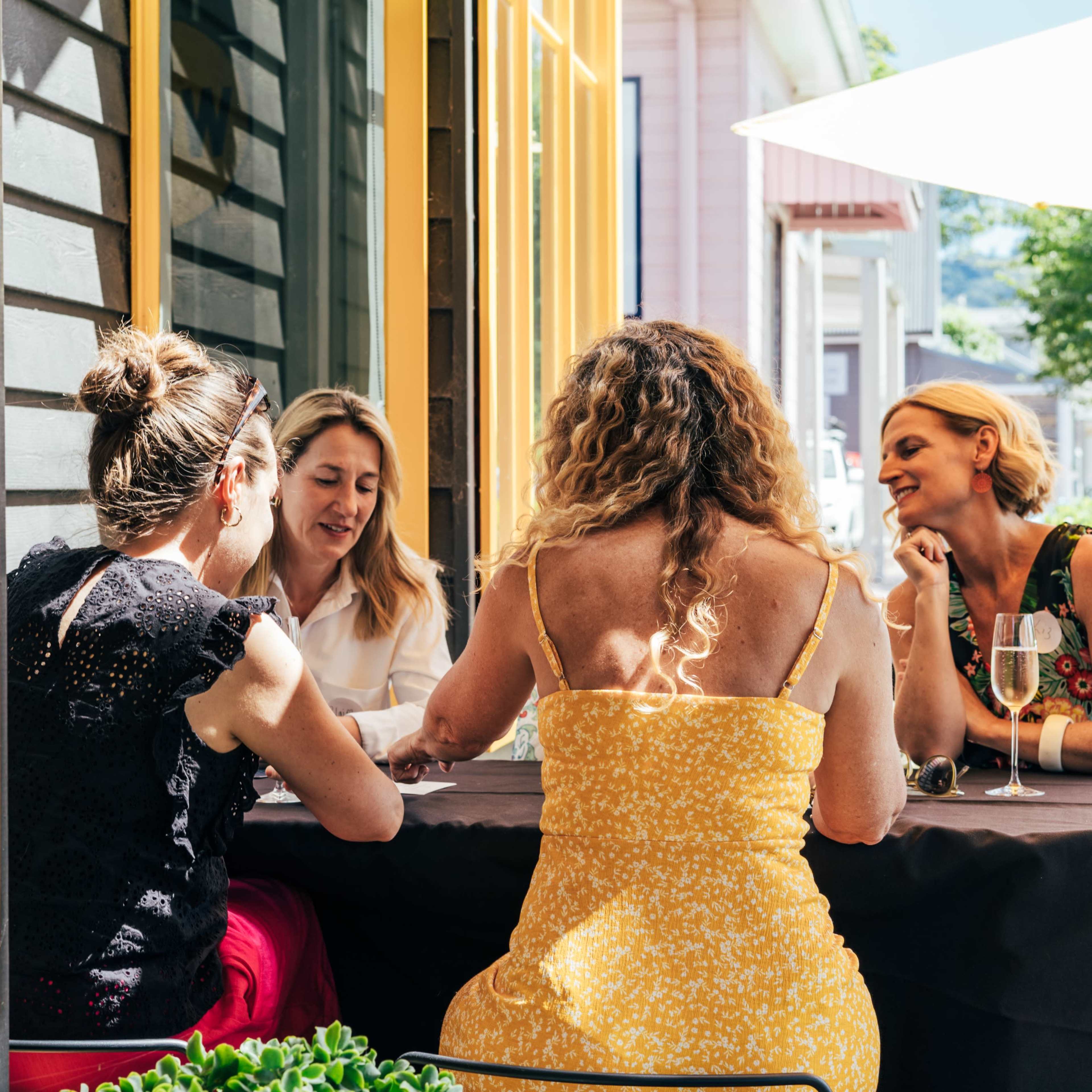 Four women sit around a table outside a café, engaged in conversation and enjoying drinks on a sunny day.
