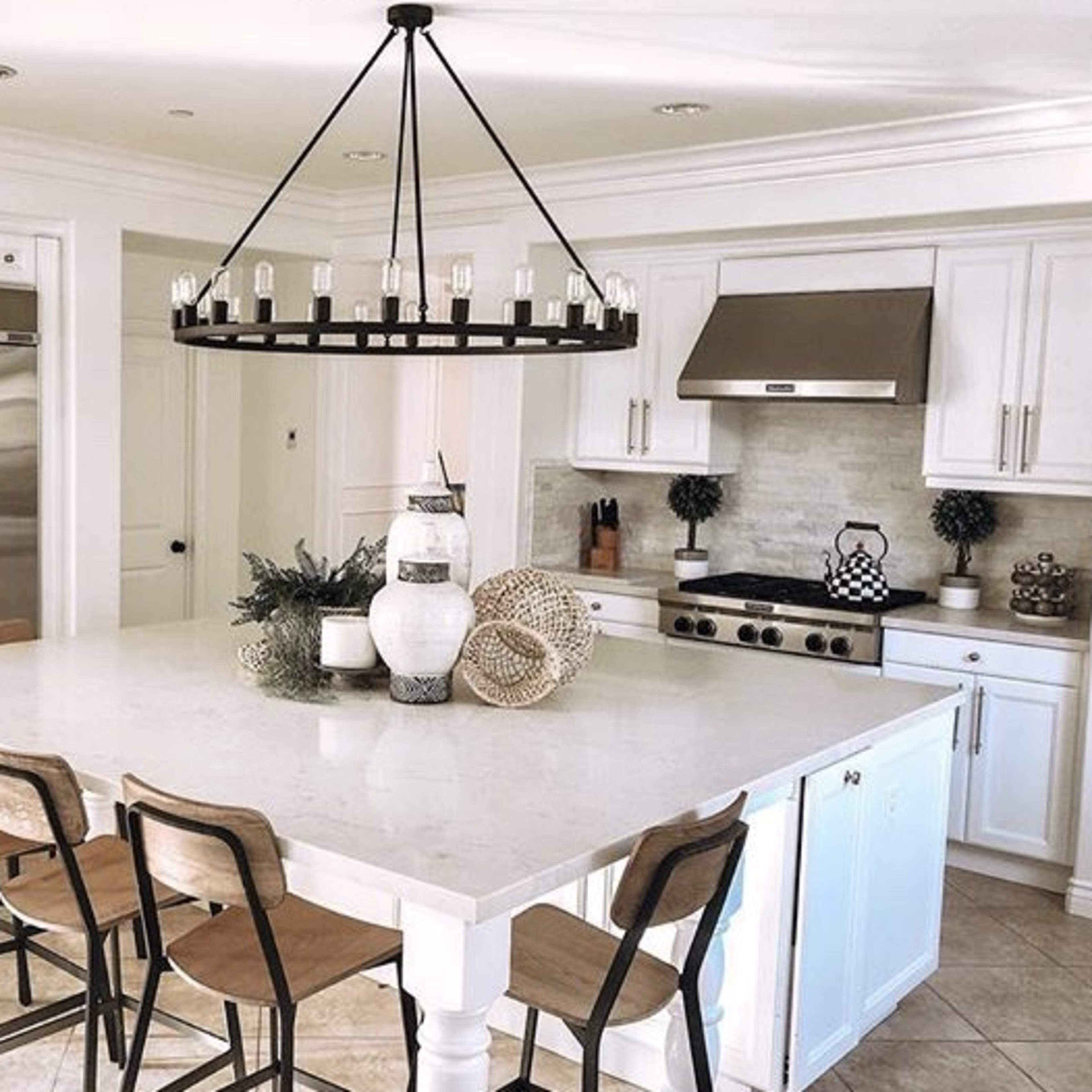 A modern kitchen features a large central island with wooden barstools, stainless steel appliances, and a circular chandelier above.