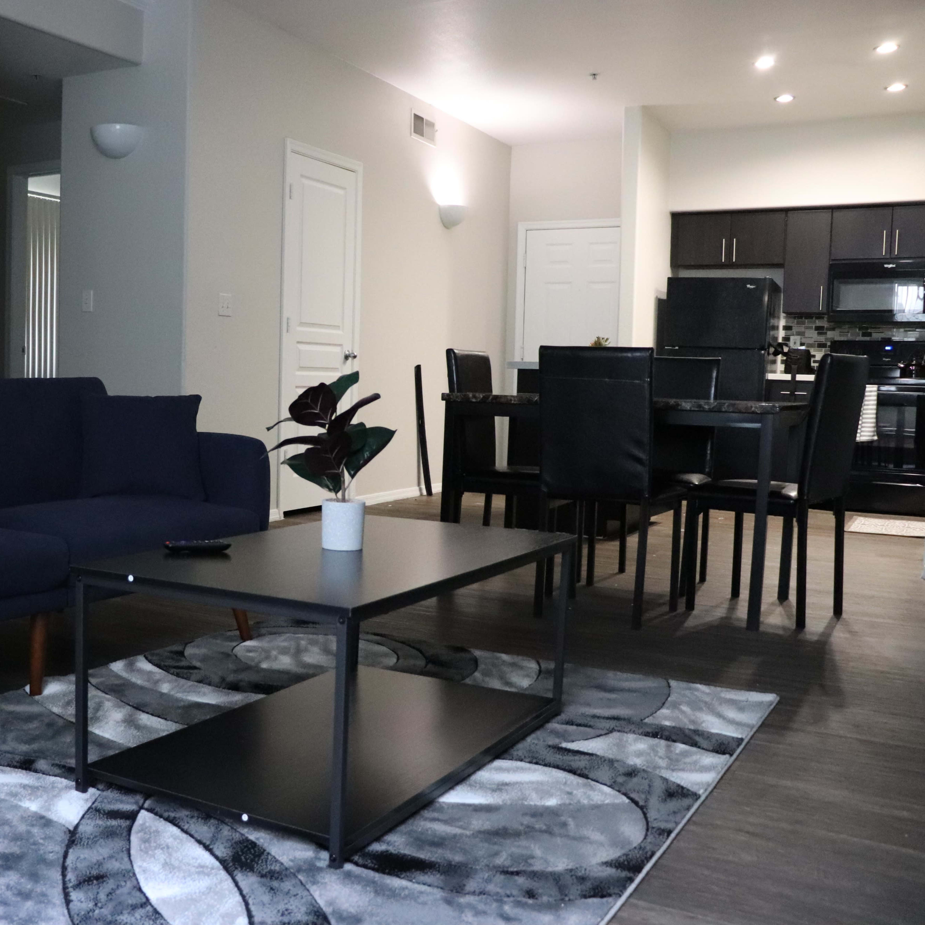 A modern living room with a blue sofa, a black coffee table on a patterned rug, and a dining area in the background.