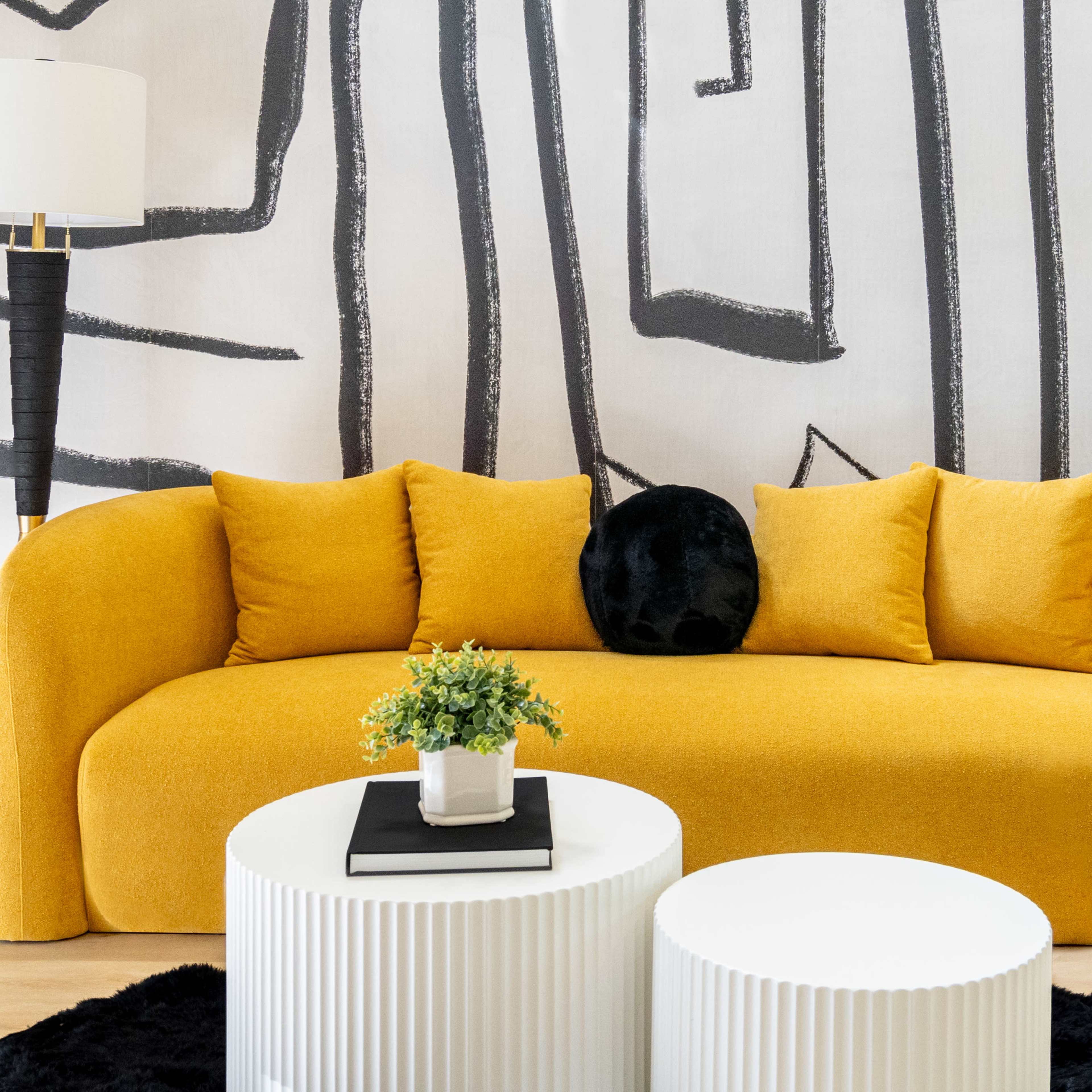 A modern, yellow curved sofa with four cushions, placed in front of a wall featuring black abstract designs, accompanied by two white round coffee tables and a small potted plant.