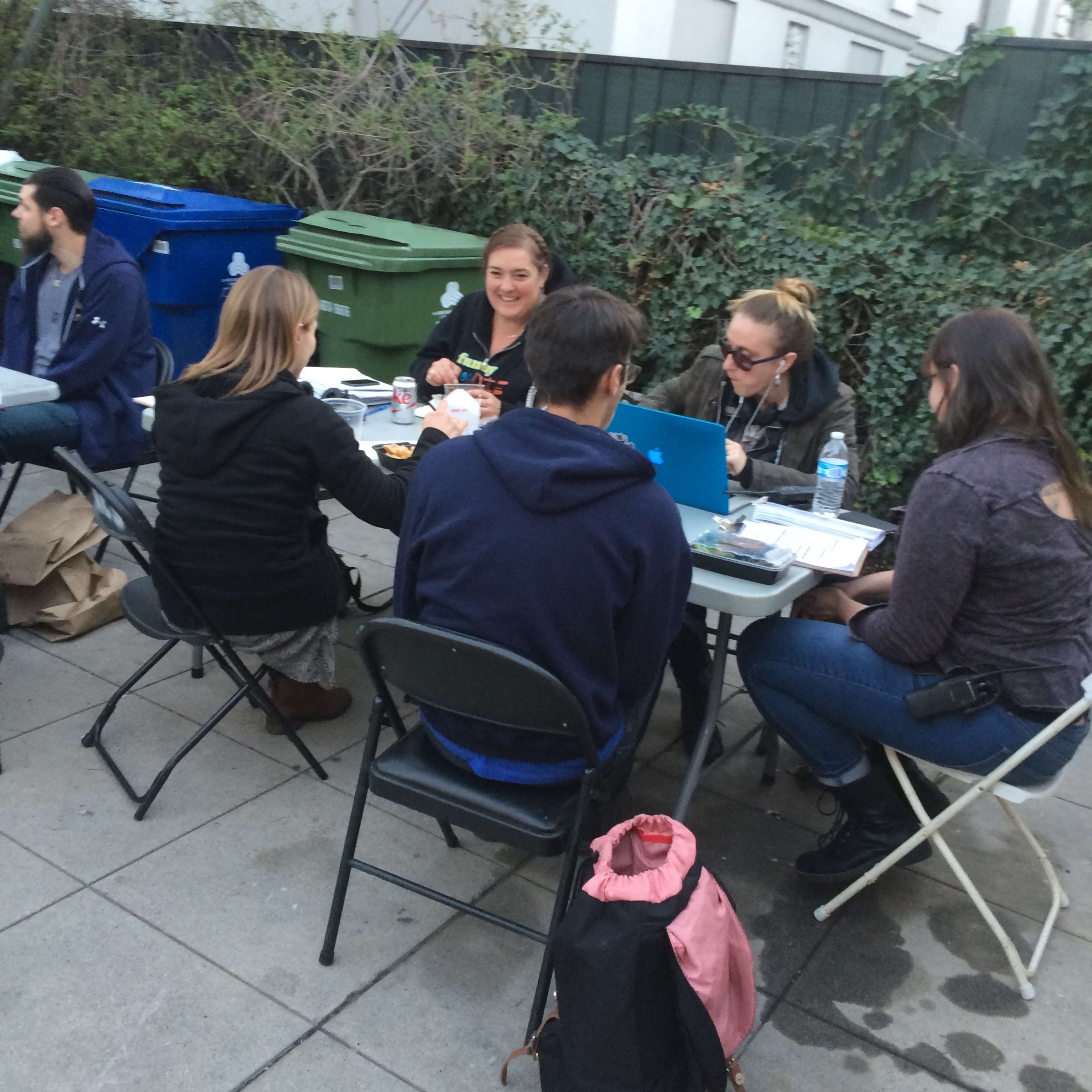A group of six people is seated around a table outdoors, engaged in work or discussion with laptops and papers spread out.
