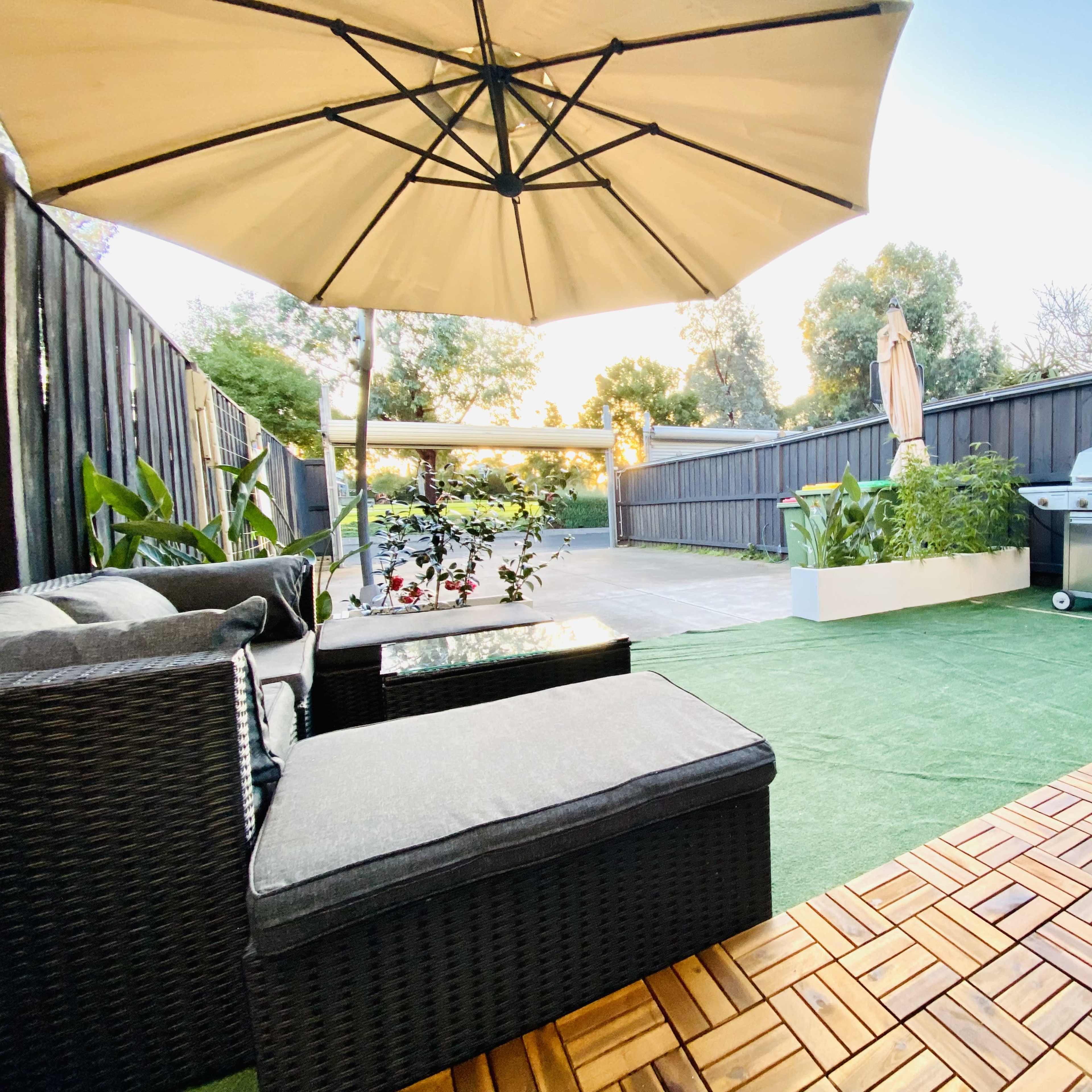 A patio area with a seating arrangement under an umbrella, artificial grass, and a barbecue grill against a wooden fence.