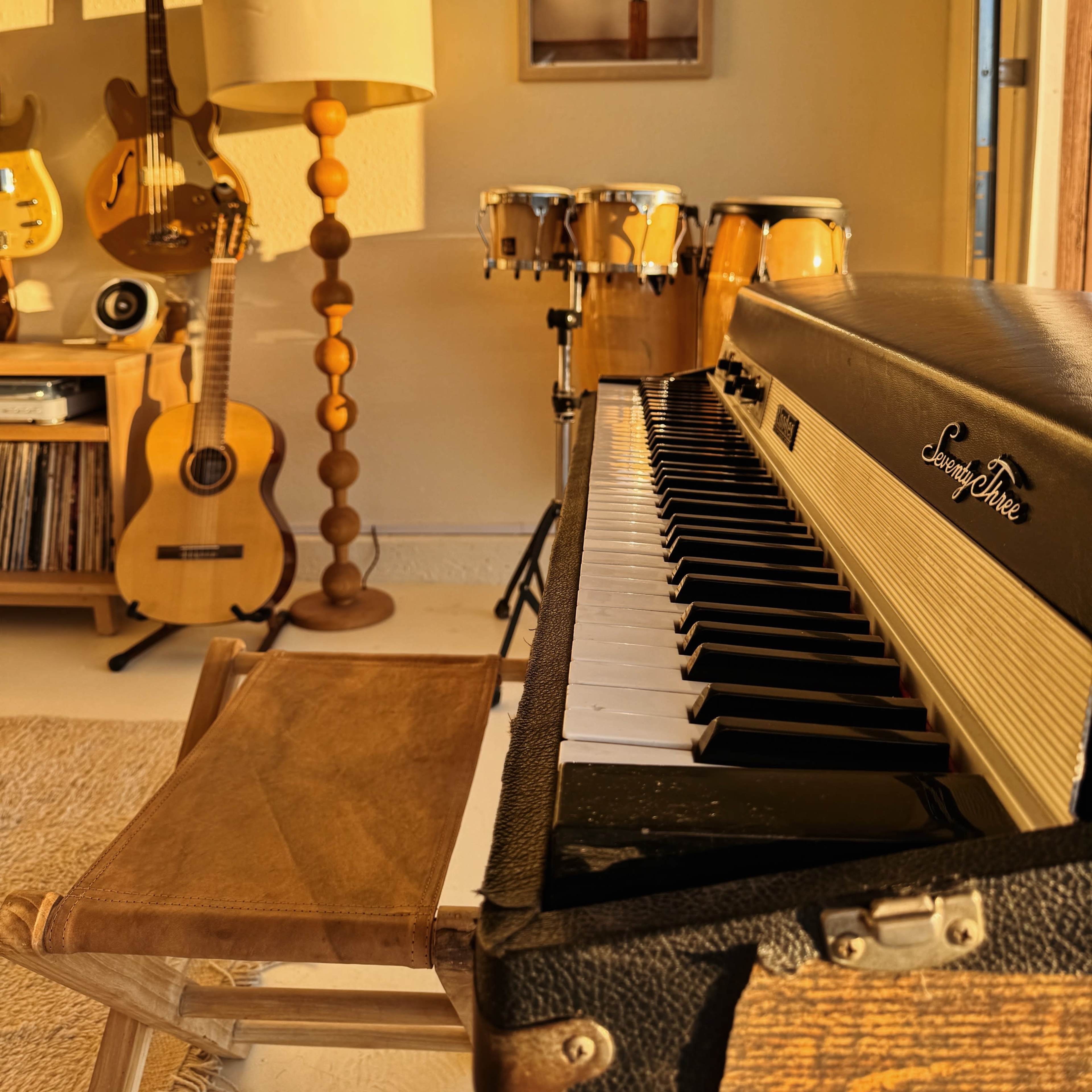 A vintage keyboard in the foreground, with various musical instruments including a lamp, a guitar, and conga drums arranged in a warm-toned interior space.