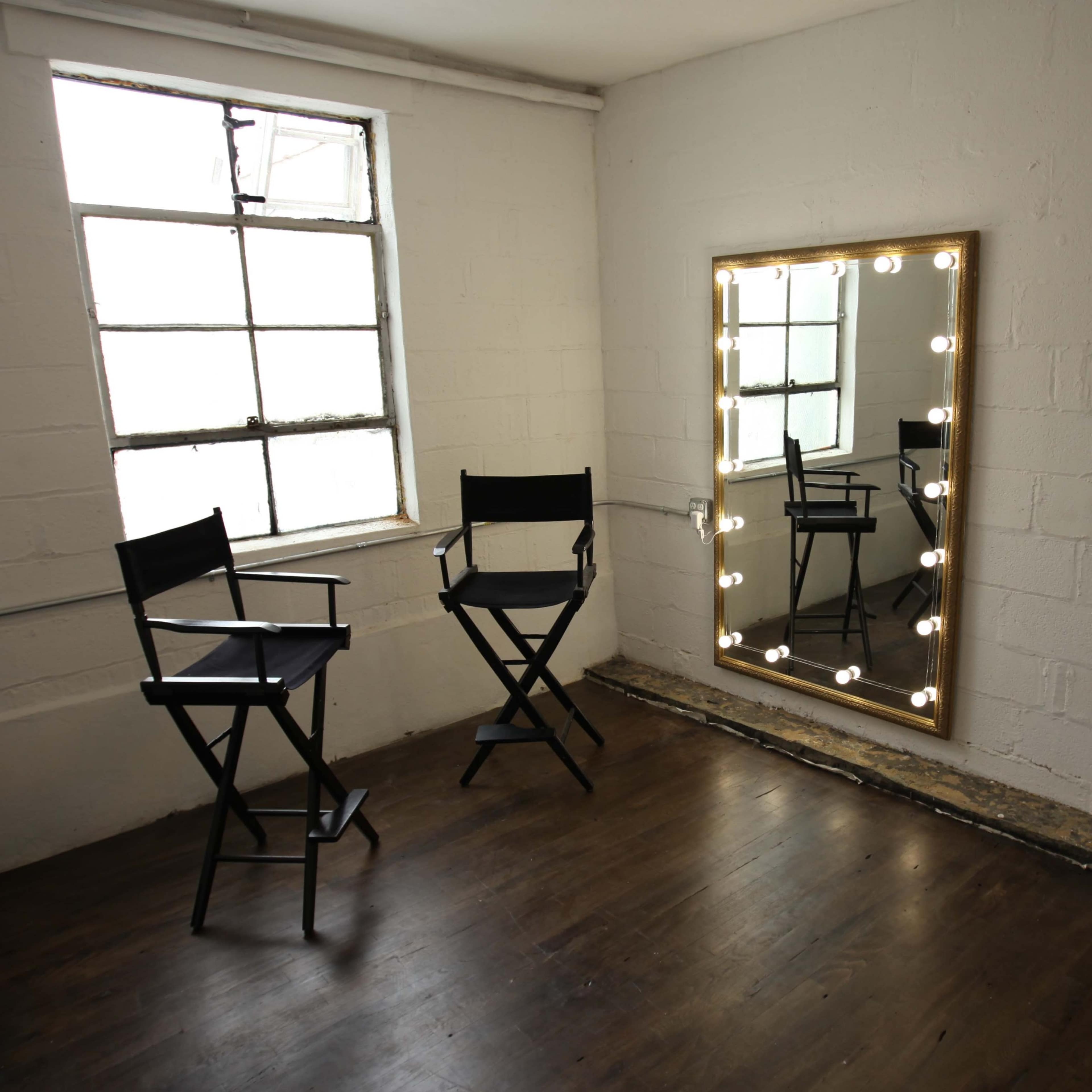 A small room featuring two director's chairs, a large illuminated mirror, and a window with multiple panes.