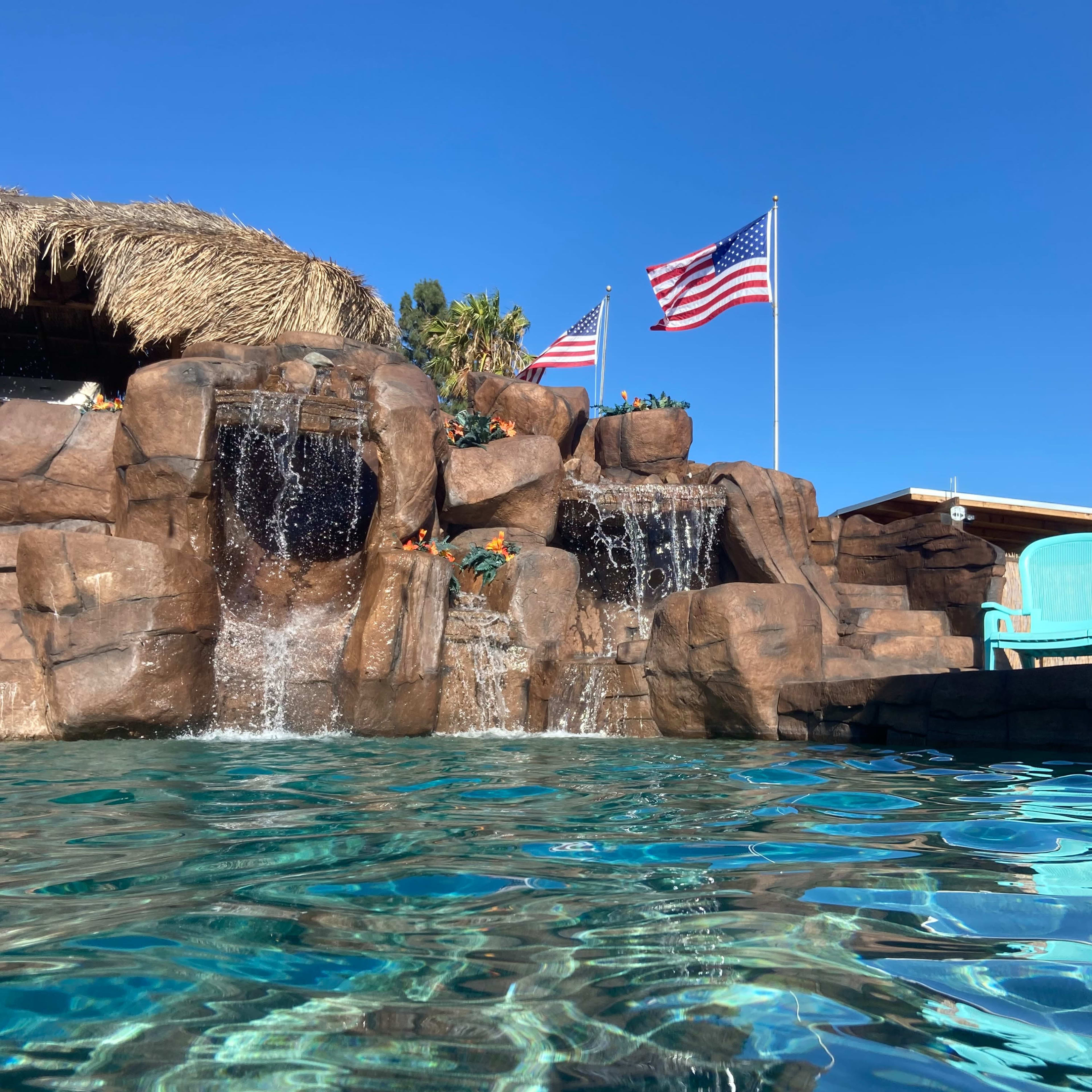 A swimming pool with a rock waterfall and two American flags waving in the background against a clear blue sky.