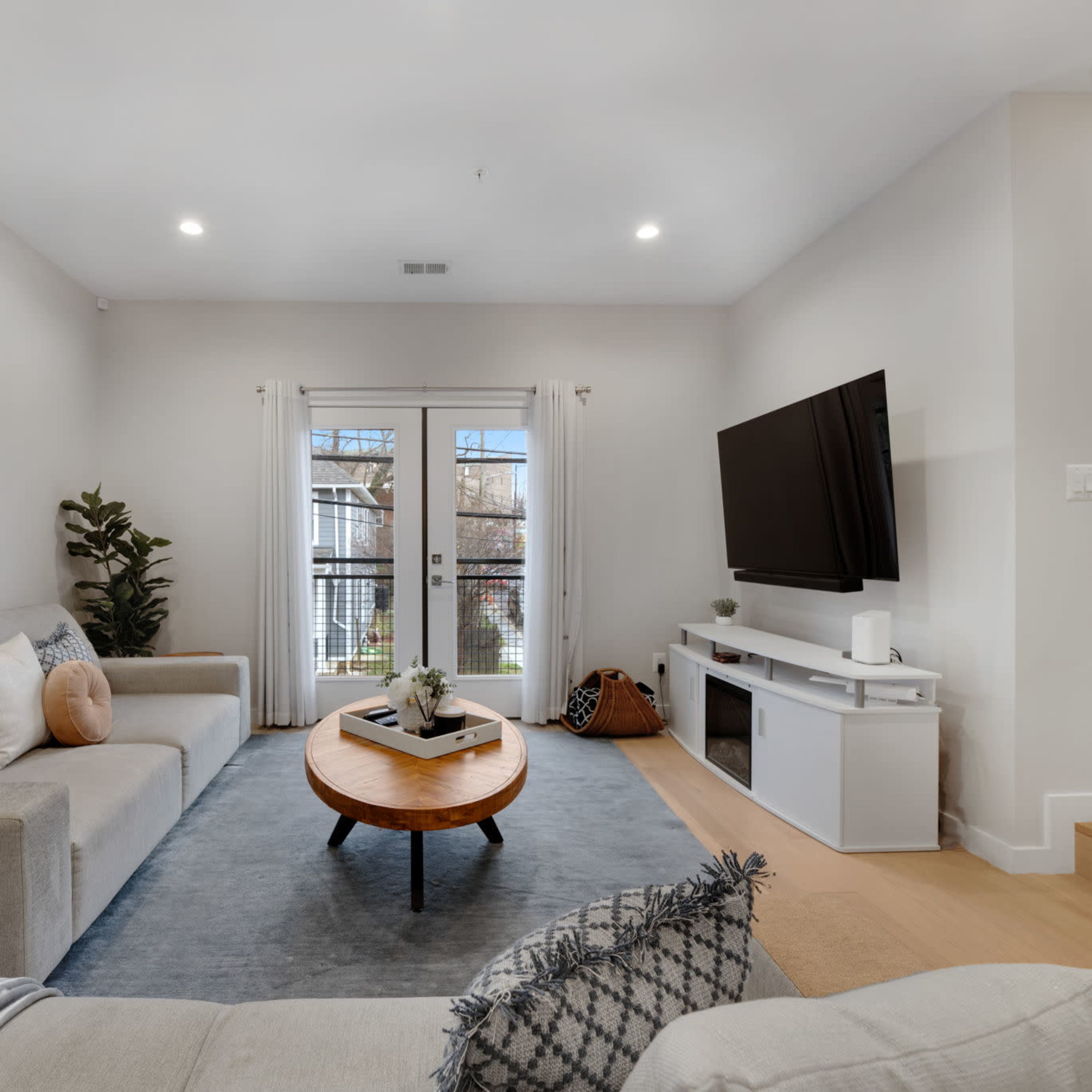 A modern living room features a light gray sectional sofa, a round coffee table, and a wall-mounted television, with large windows overlooking a balcony.