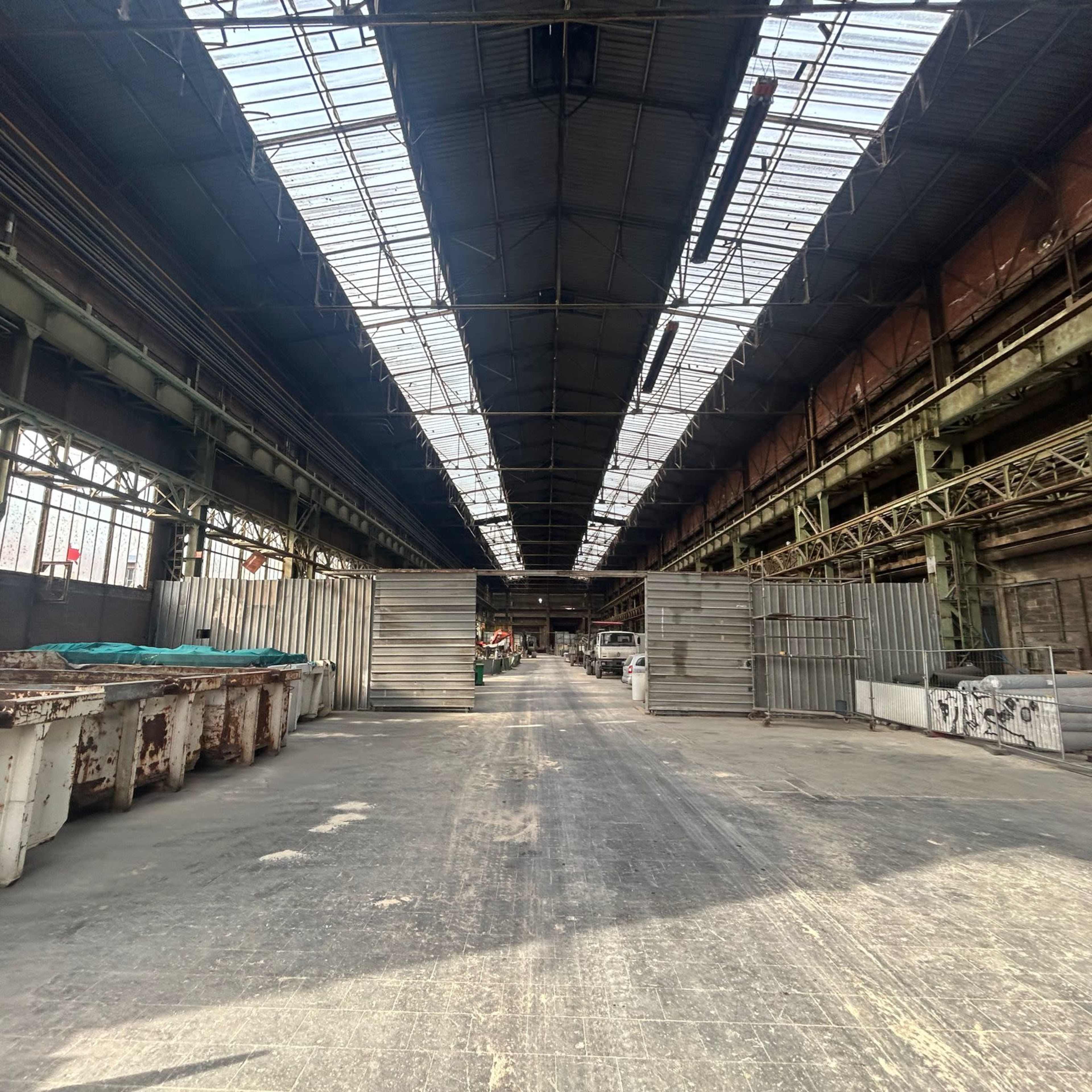 A long, industrial warehouse interior with a clear path running down the center, flanked by metal containers and large overhead windows allowing natural light to enter.