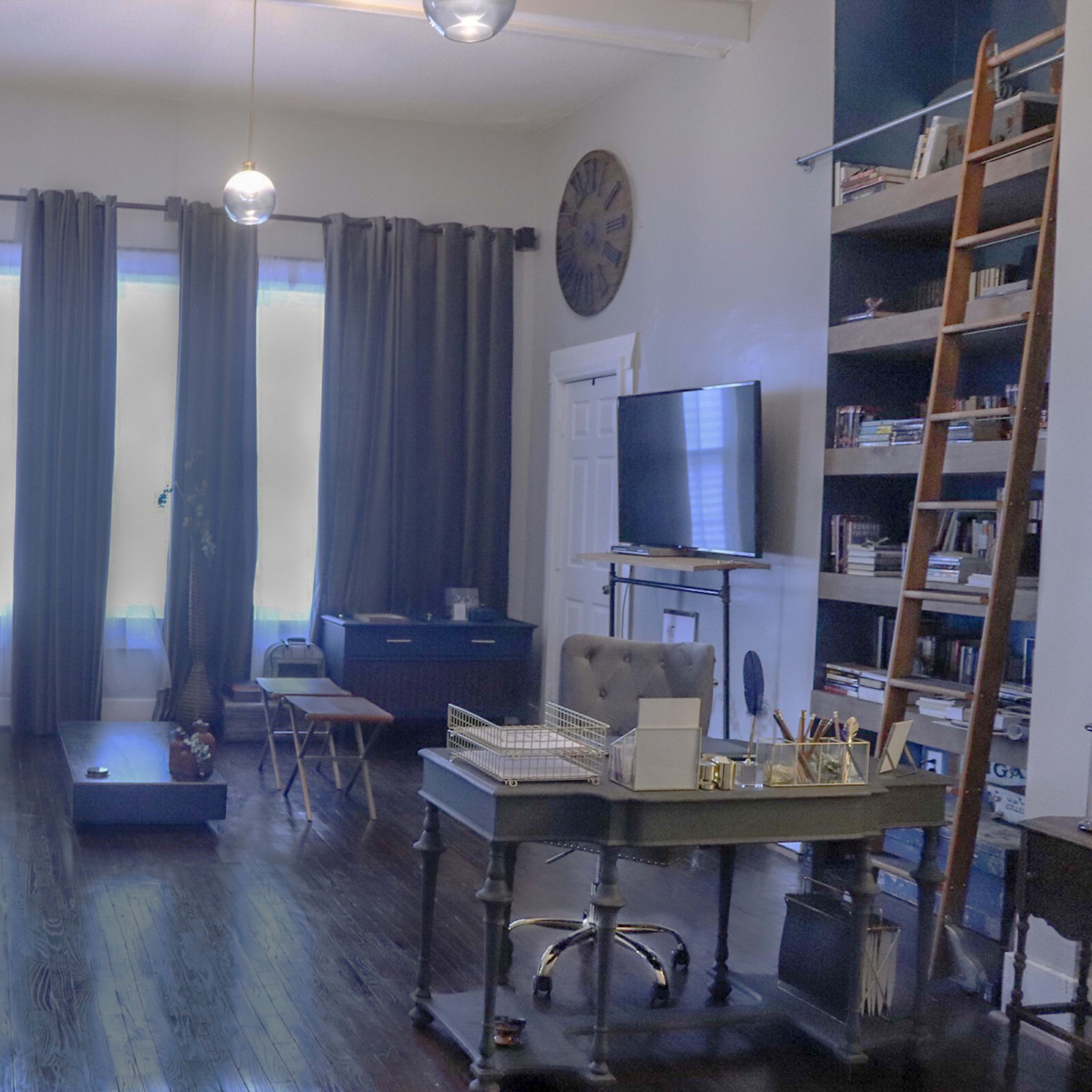 A spacious living room features a blue sofa, a wooden coffee table, and a study desk against a backdrop of large windows with dark curtains.