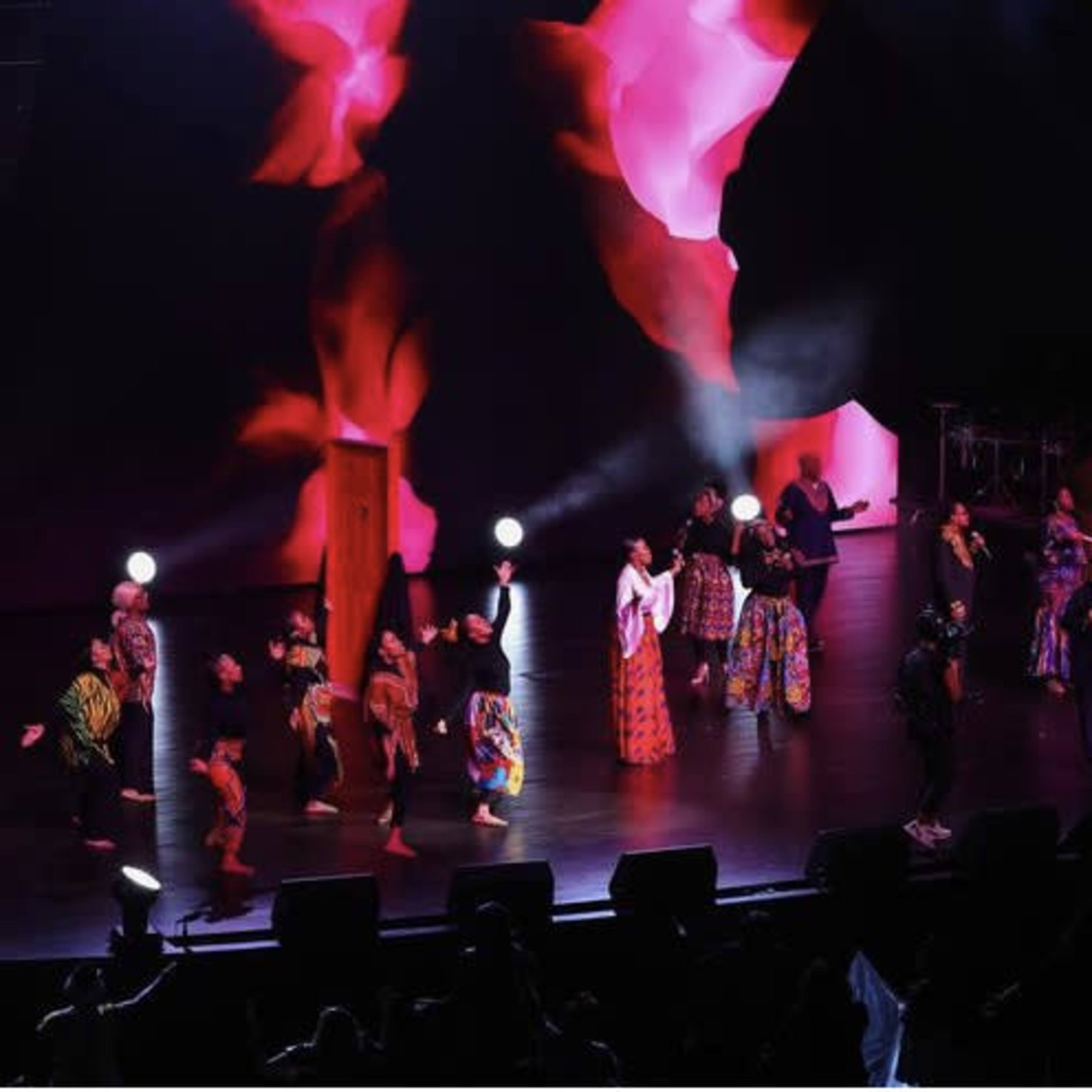 A group of performers with colorful costumes is dancing on stage under vibrant lighting during a live event.