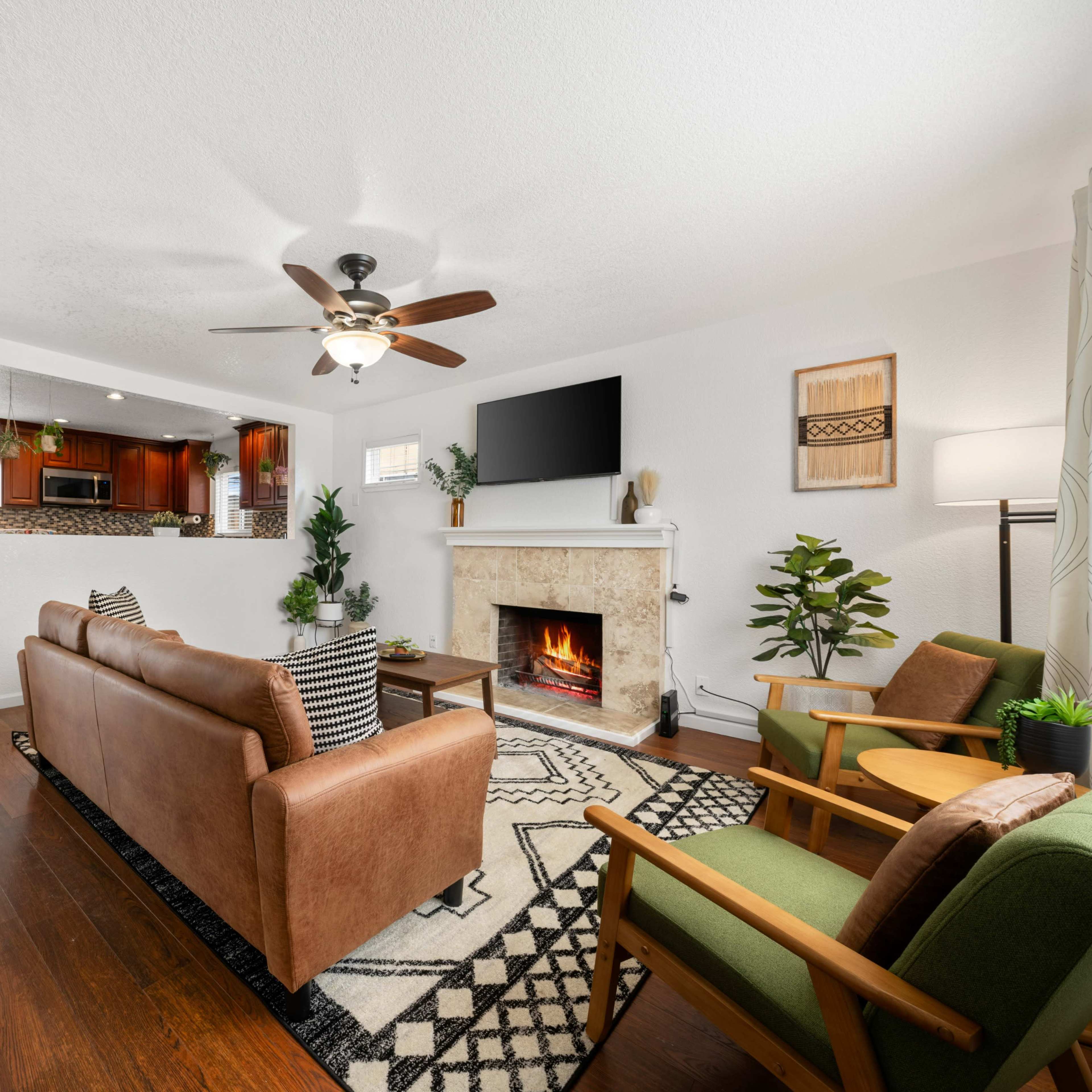 A modern living room featuring a brown leather sofa, two green armchairs, a fireplace, and a view of a kitchen in the background.
