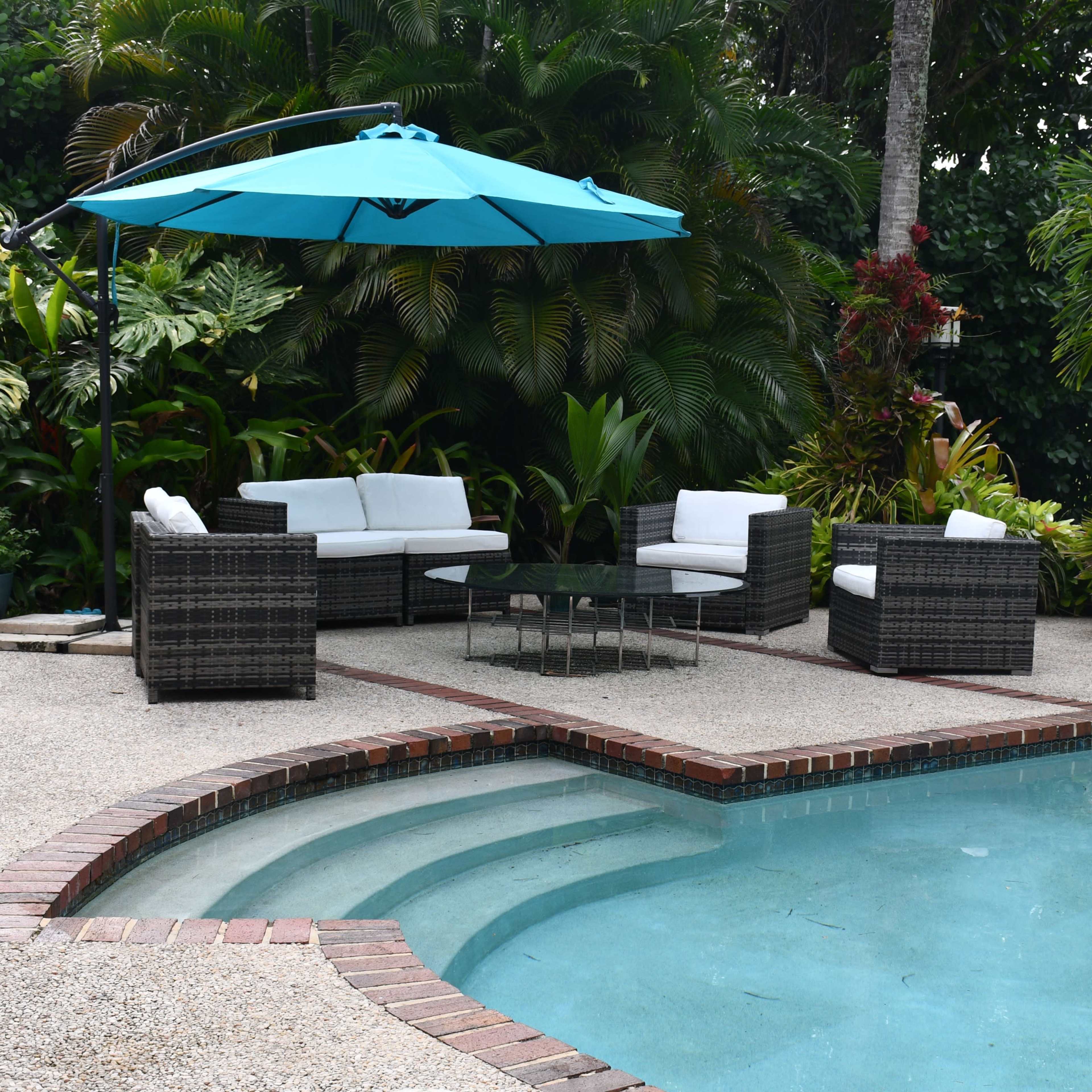 A patio with a turquoise umbrella shades a seating area near a shaped swimming pool surrounded by tropical plants.