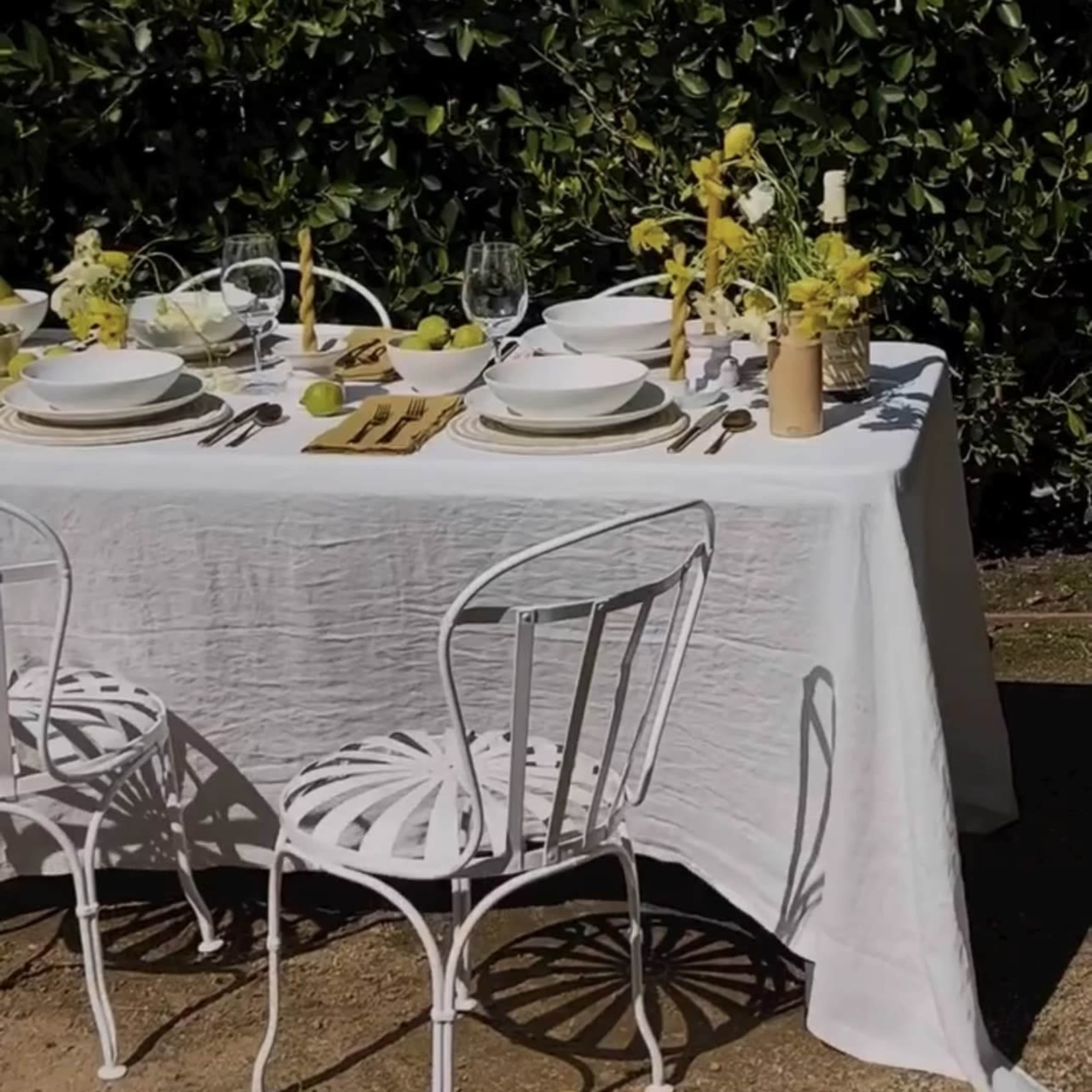 A table is set for dining, covered with a white tablecloth, featuring plates, glasses, and decorative elements, surrounded by white chairs in a garden.