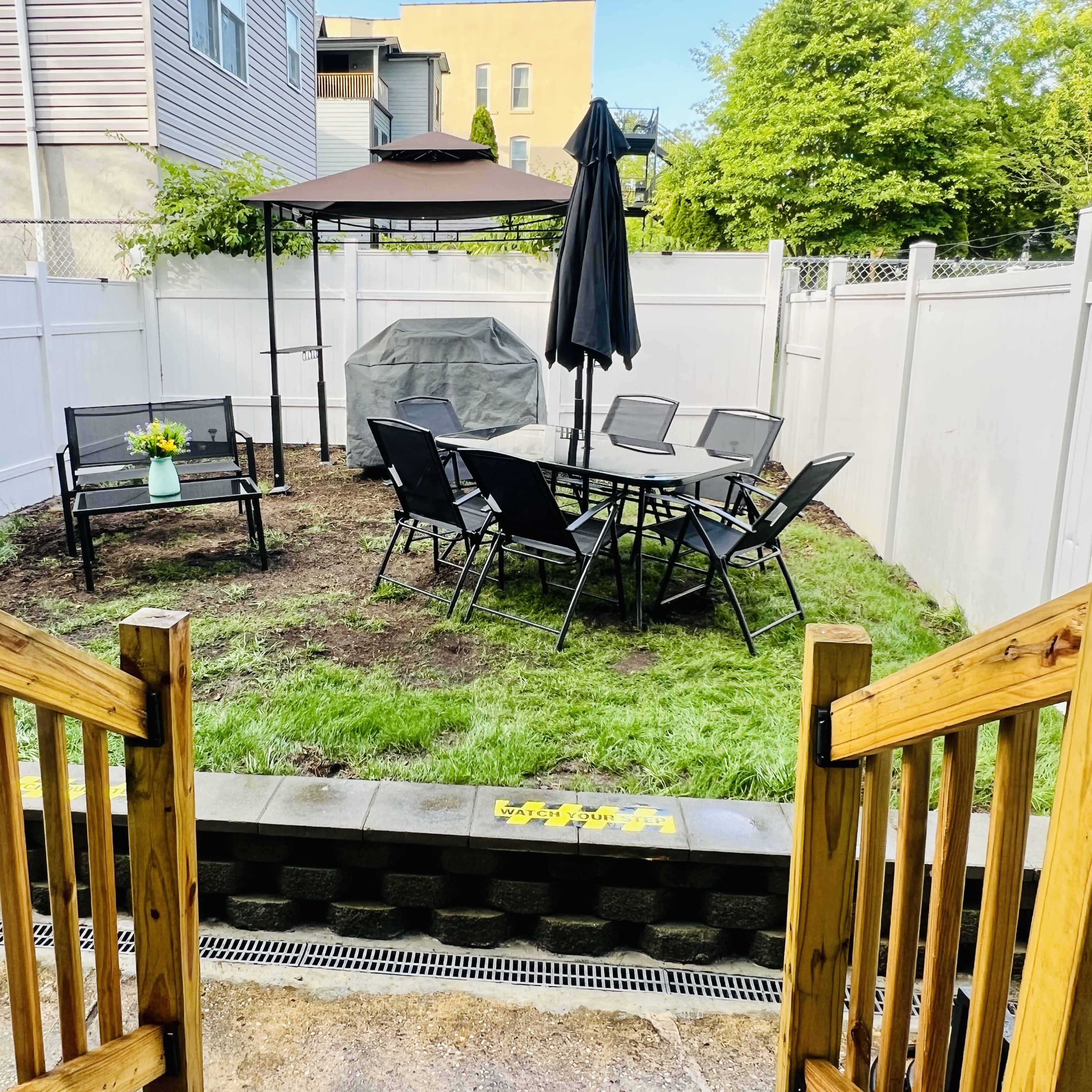A backyard space features a seating area with chairs and a table, a shaded gazebo, and a grassy lawn enclosed by a white fence.