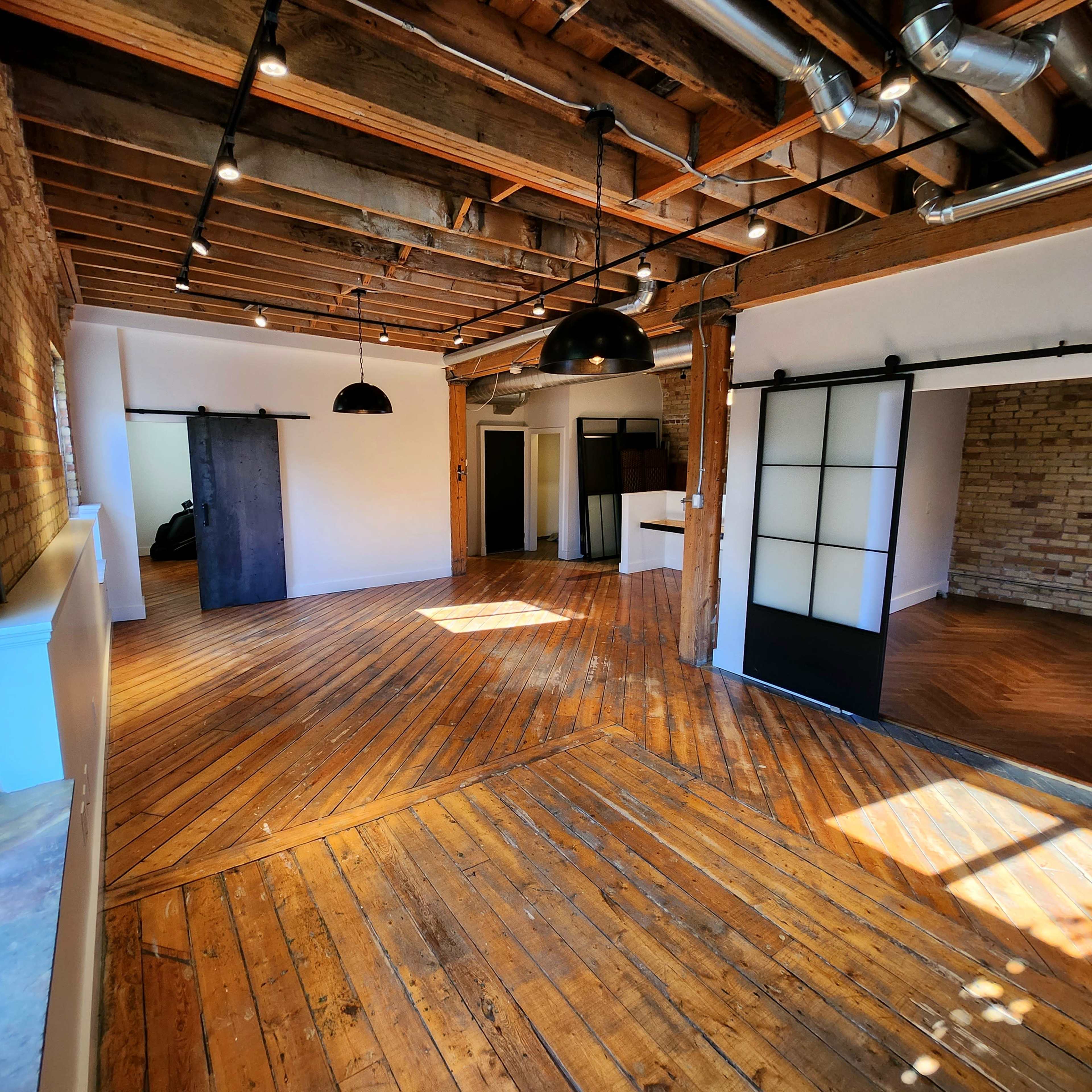 A spacious room with wooden floors, exposed beams, and large windows, featuring a minimalist design with a sliding door and pendant lighting.
