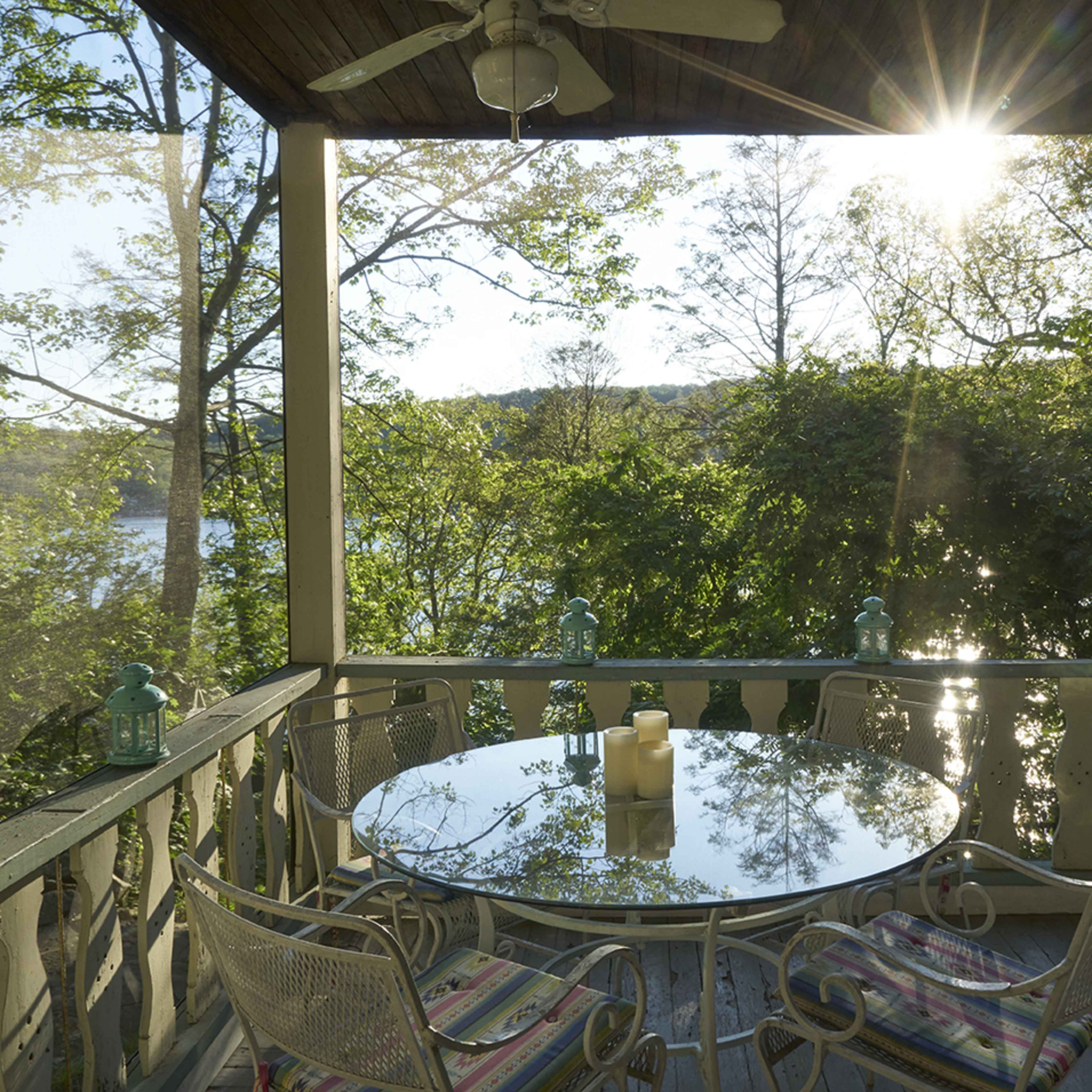 A round glass table with metal chairs is set on a porch overlooking a river framed by trees, with sunlight streaming through the foliage.