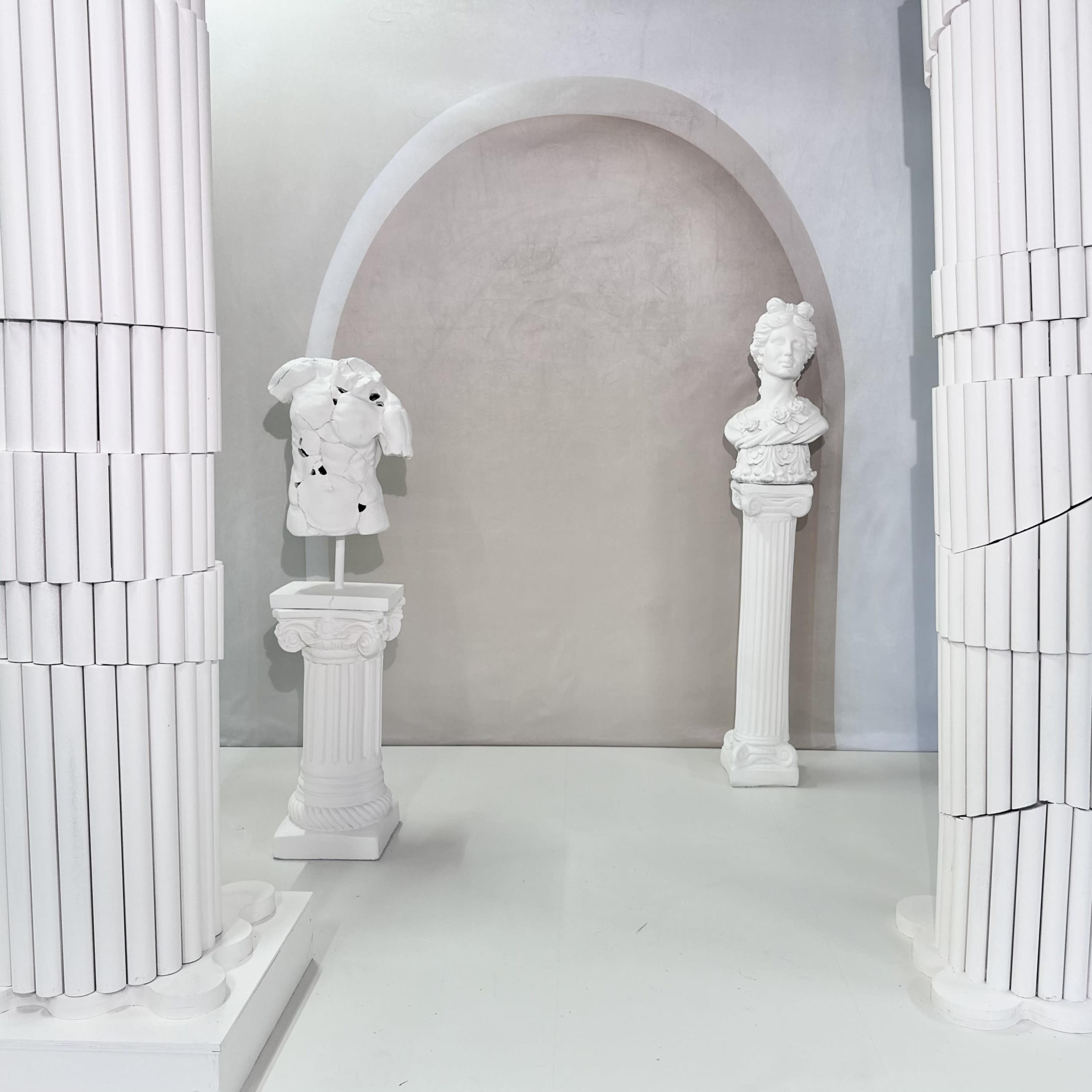 A minimalist display of classical white sculptures and columns arranged within an arched backdrop.
