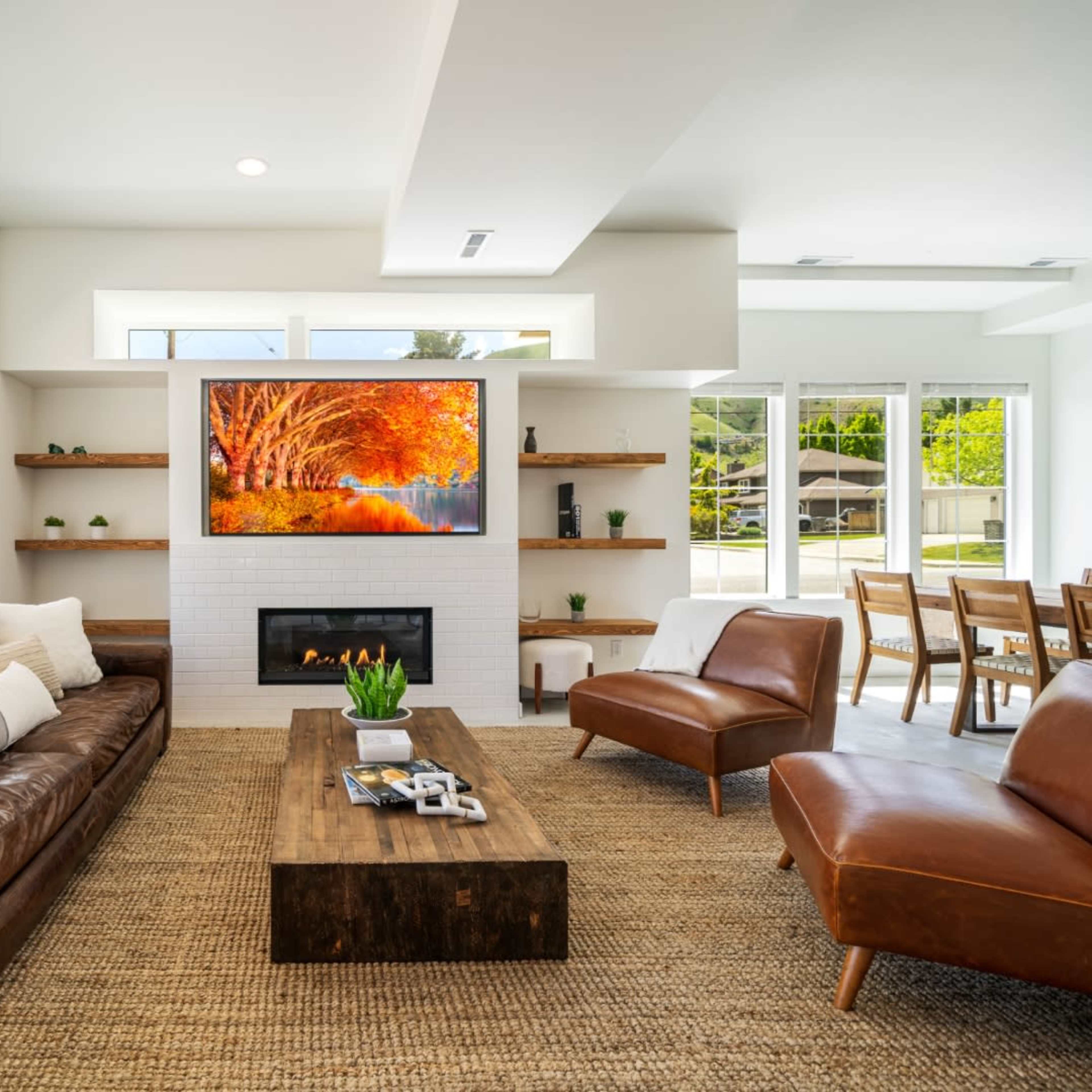 A modern living room features a brown leather sofa, a wooden coffee table, and a large wall-mounted television displaying an autumn scene.