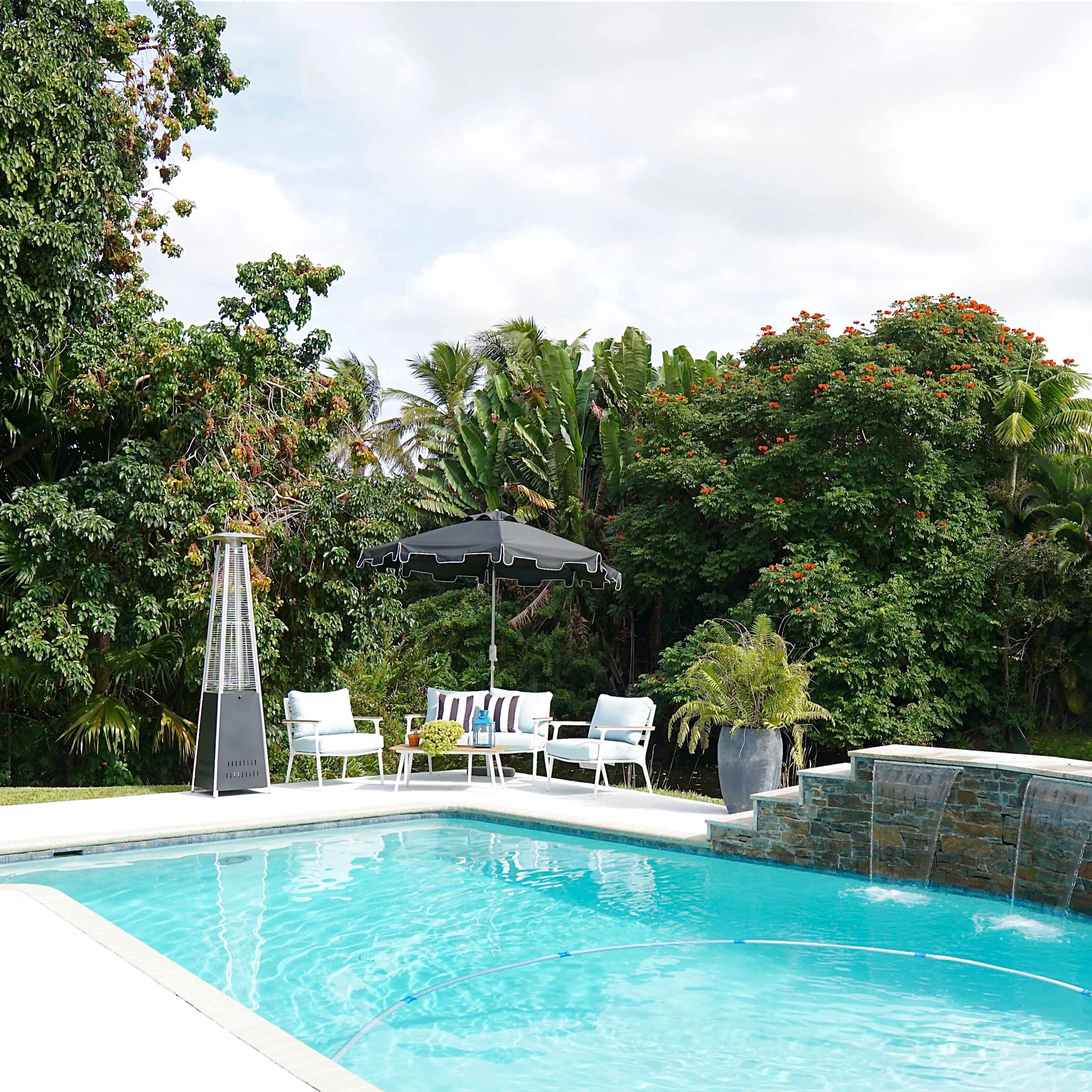 A clear blue swimming pool is surrounded by lush greenery and a patio with white chairs and a black umbrella.