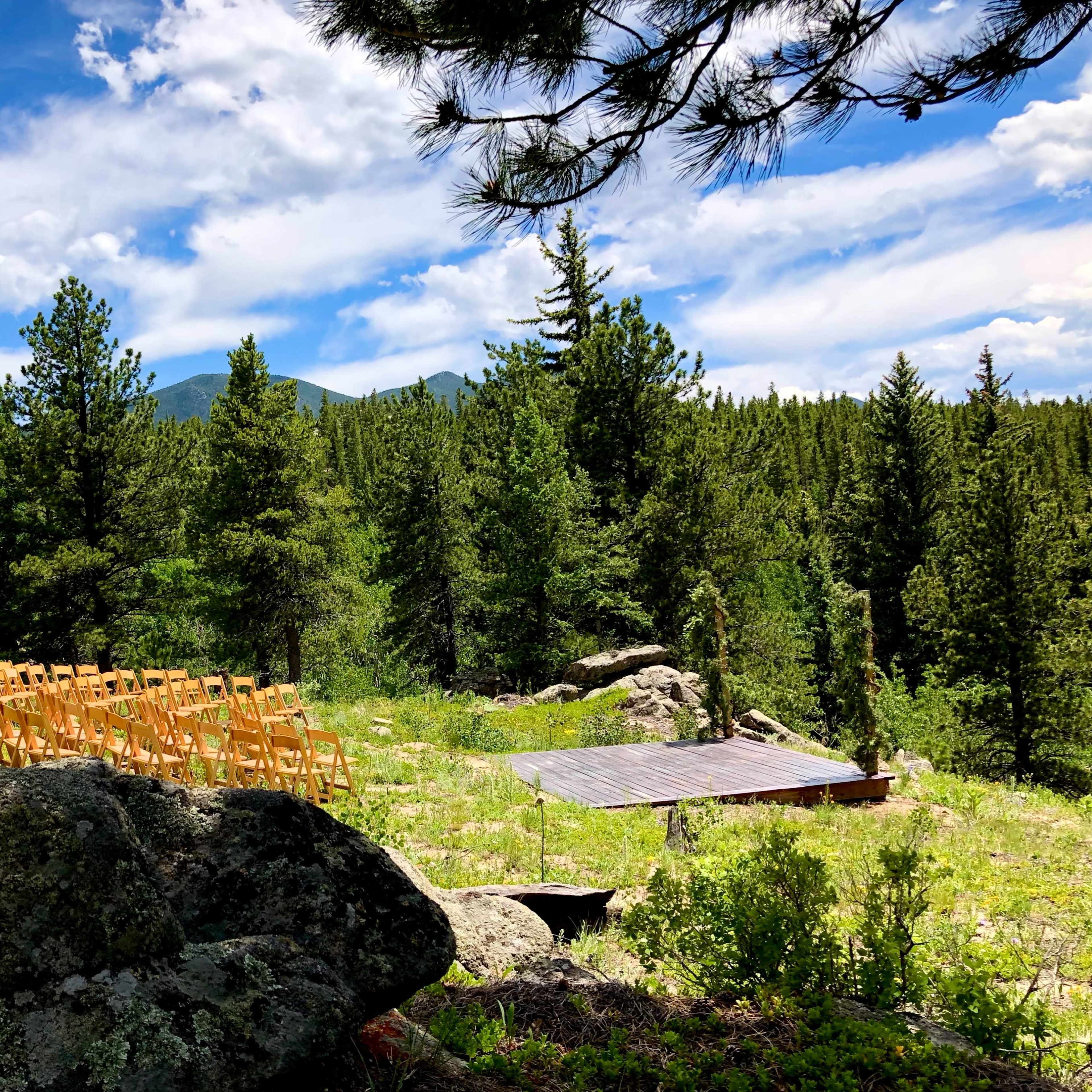 A wooden platform stands in a clearing surrounded by trees, with rows of empty chairs set up for an event.