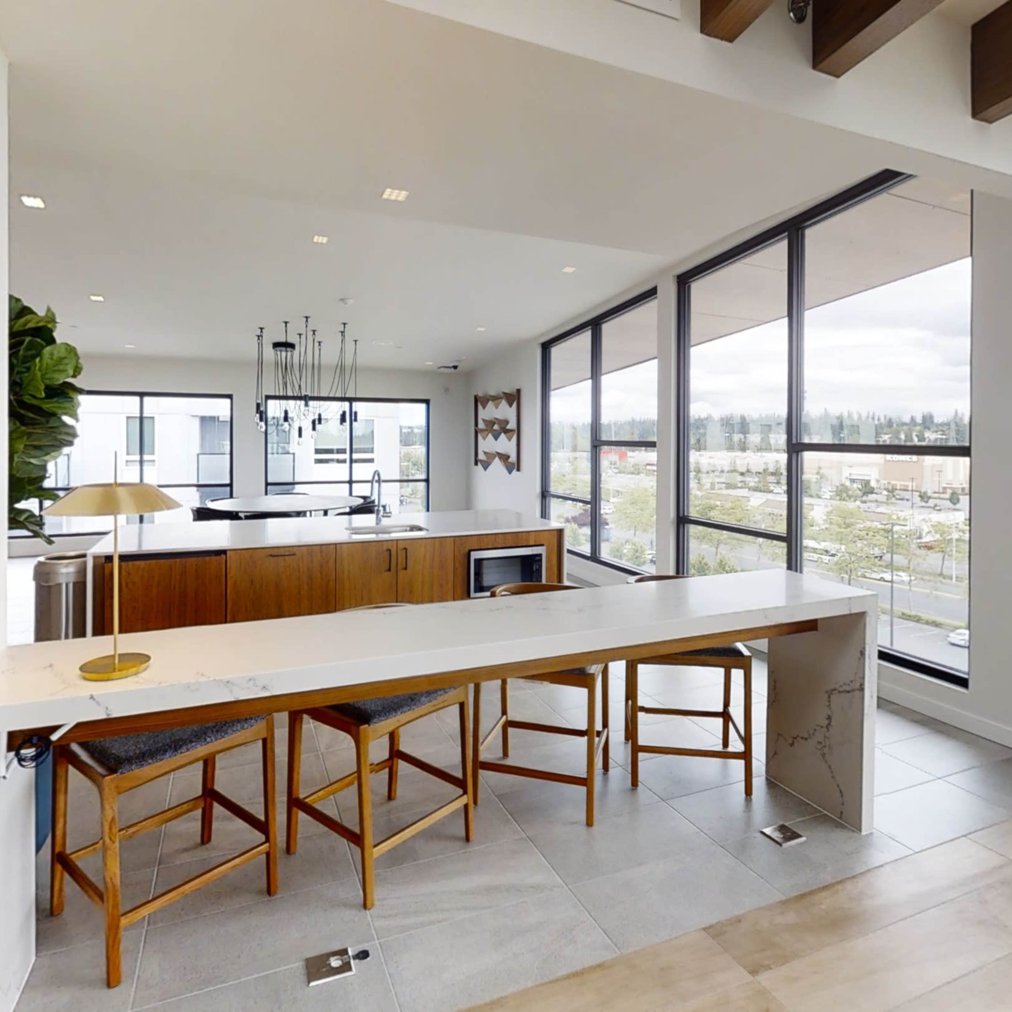 A modern kitchen with a countertop and three wooden stools, featuring large windows that provide a view of the exterior landscape.