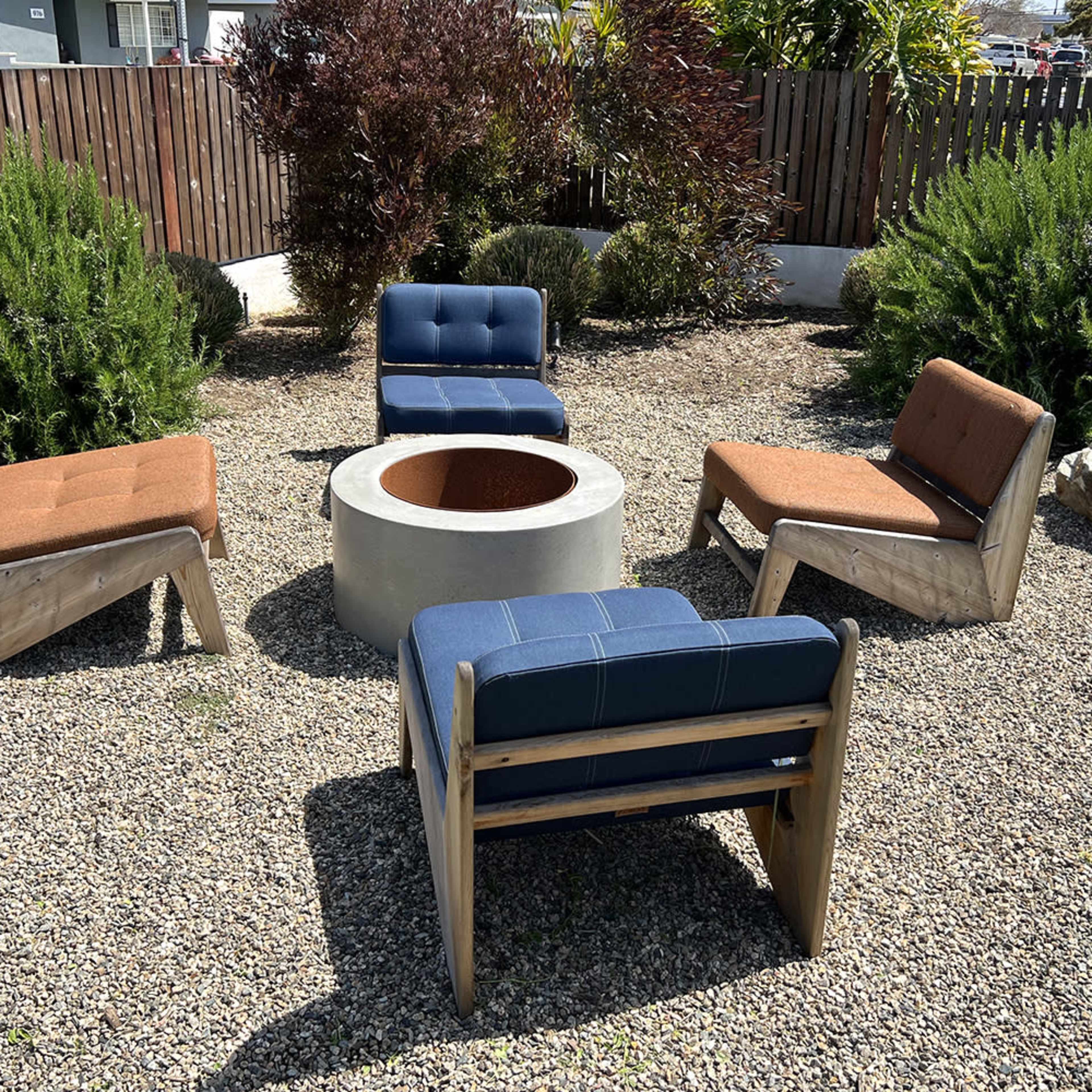 A circular stone fire pit is surrounded by four modern chairs on a gravel surface in a landscaped area.