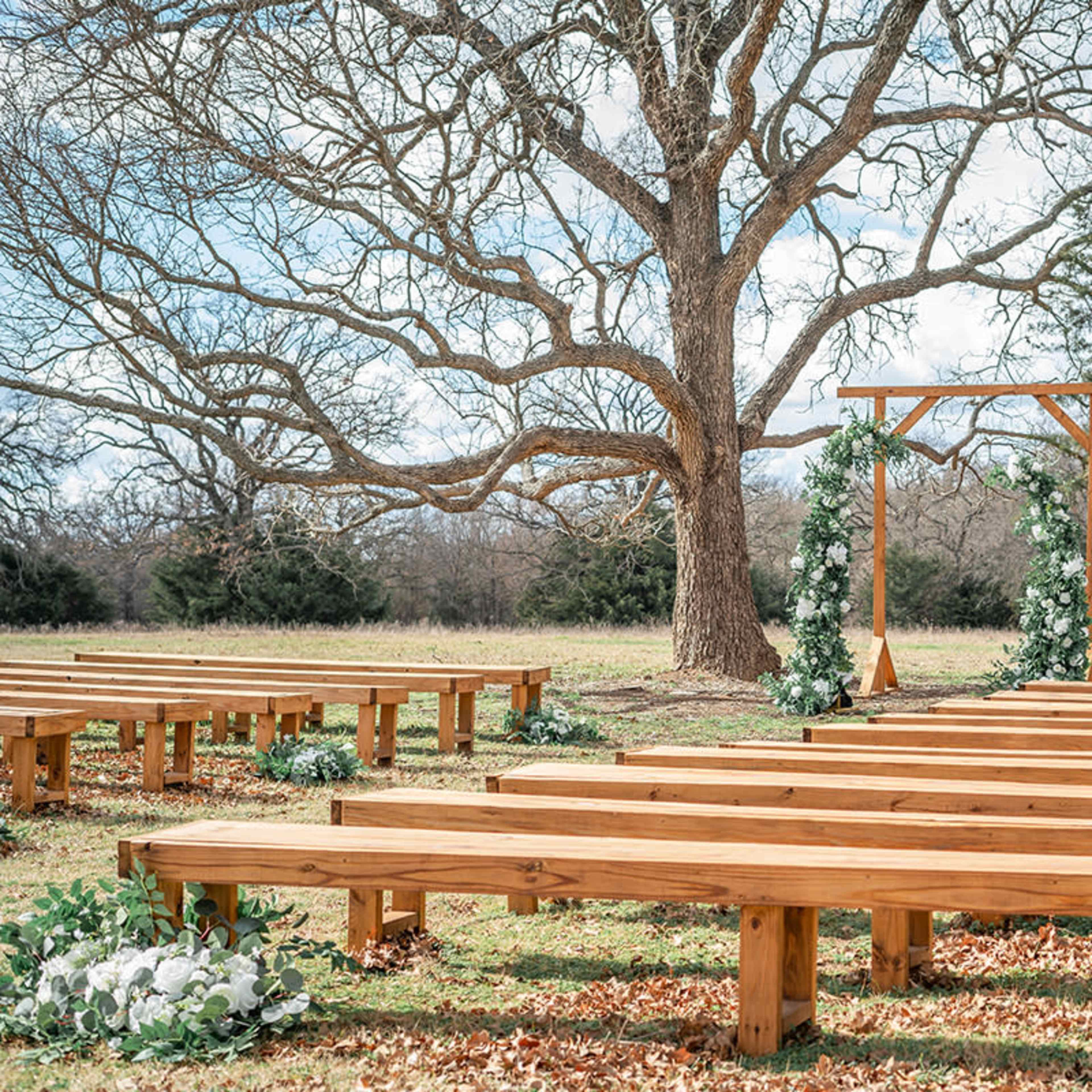 A wooden outdoor wedding ceremony setup features benches arranged in rows facing a decorated arch under a large tree.