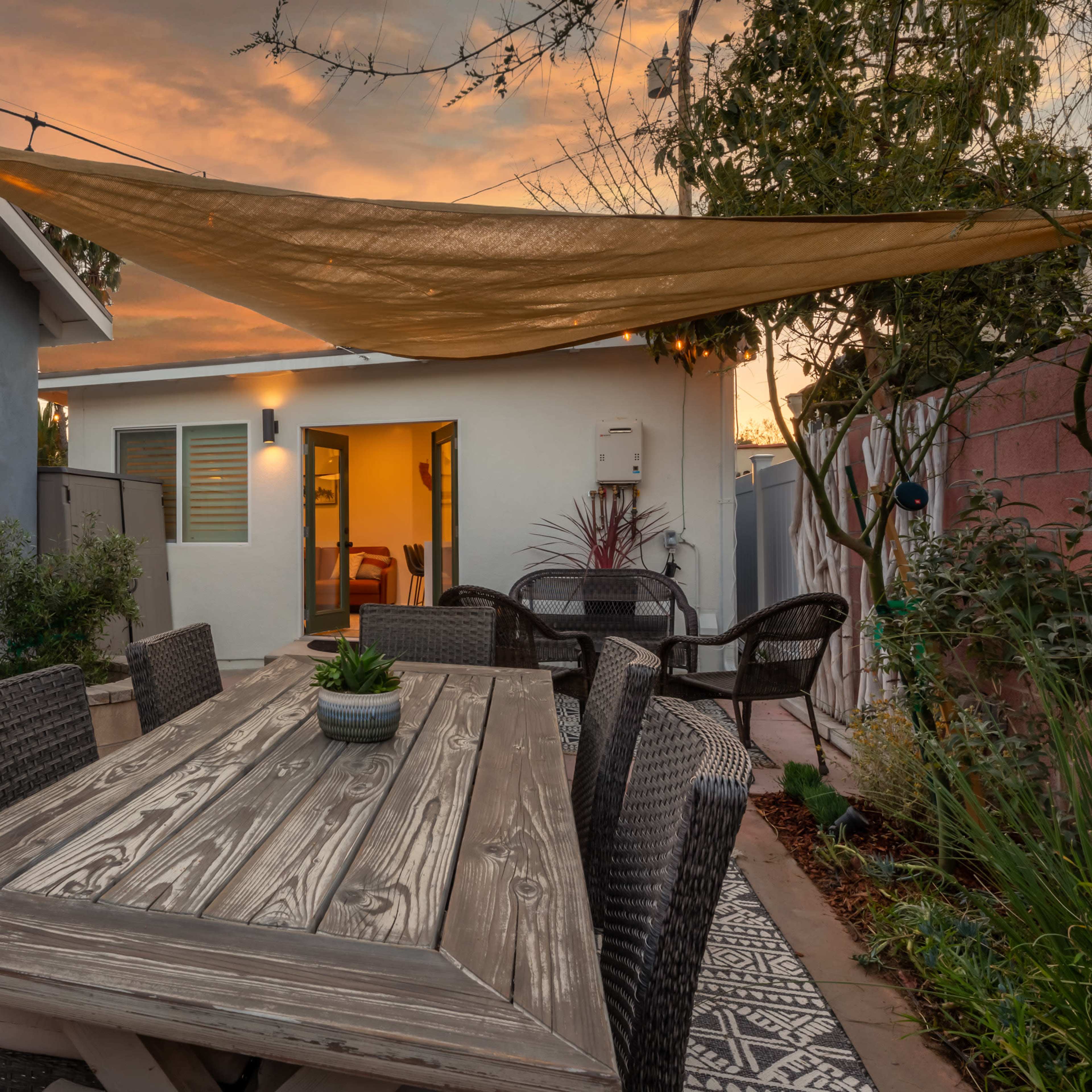 A wooden dining table with wicker chairs is set under a canopy in a patio area, surrounded by plants and a house in the background at sunset.
