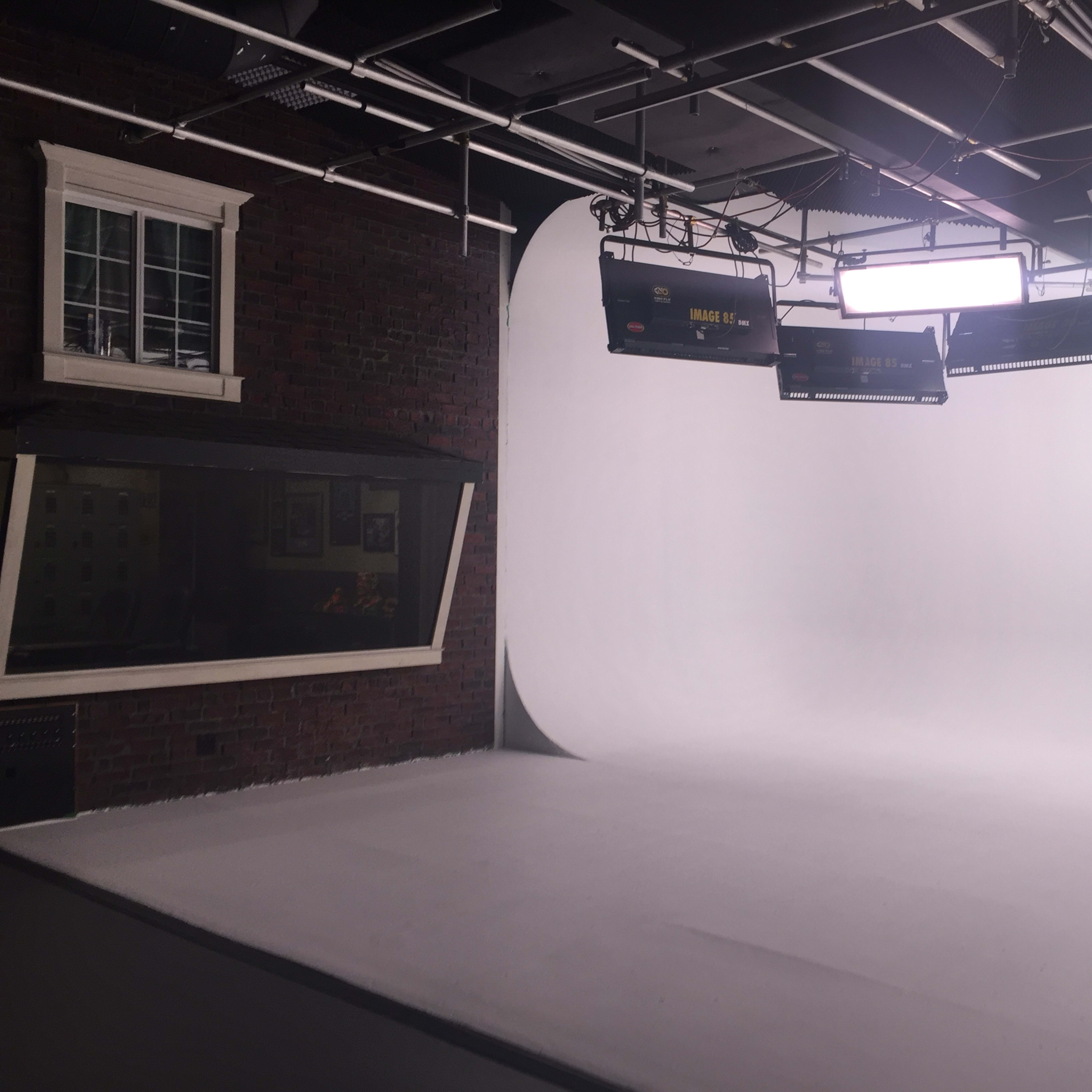 A photography studio with a plain white backdrop, studio lights hanging from the ceiling, and a brick wall to one side.