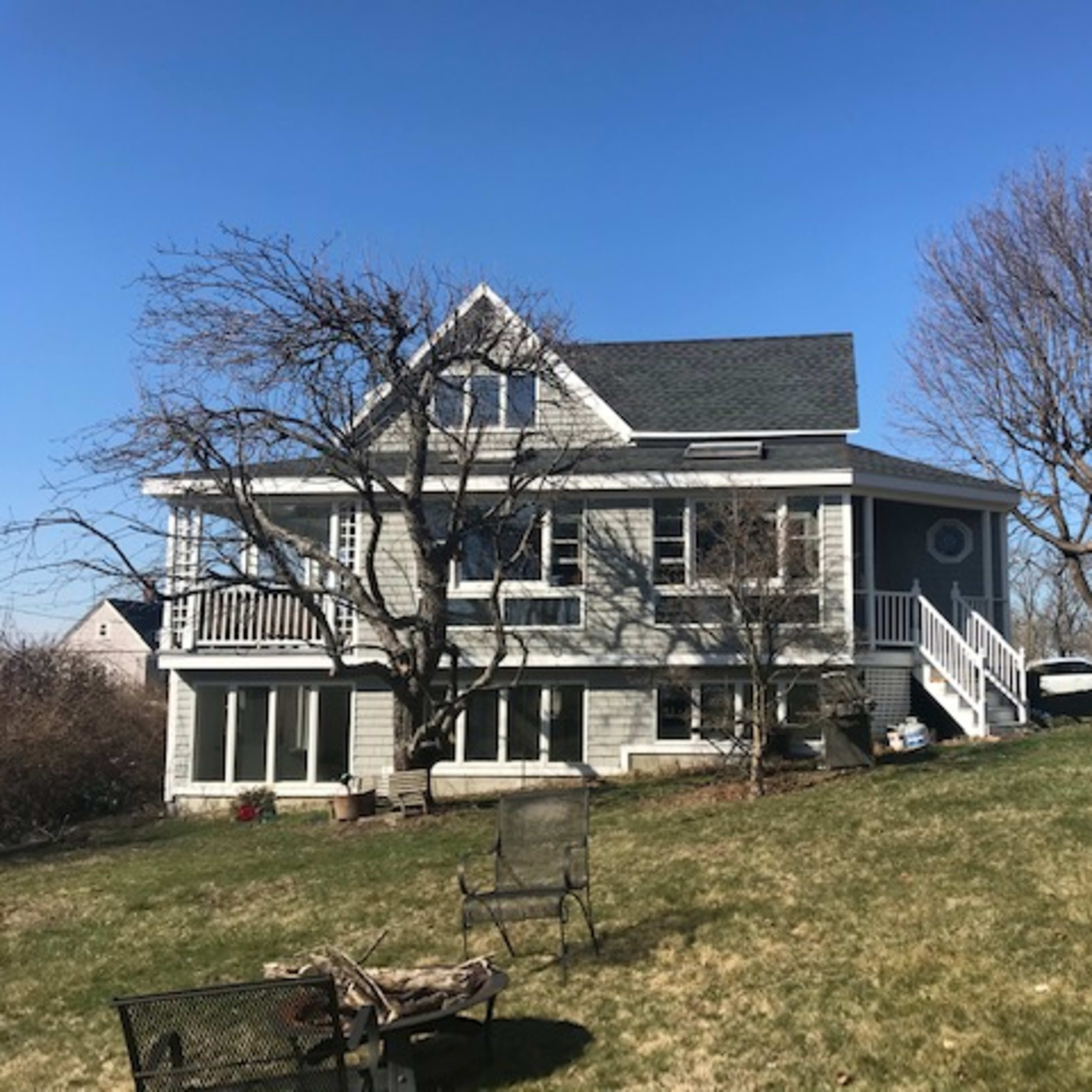 A two-story house with a gray exterior and a large yard featuring bare trees and a few outdoor chairs.