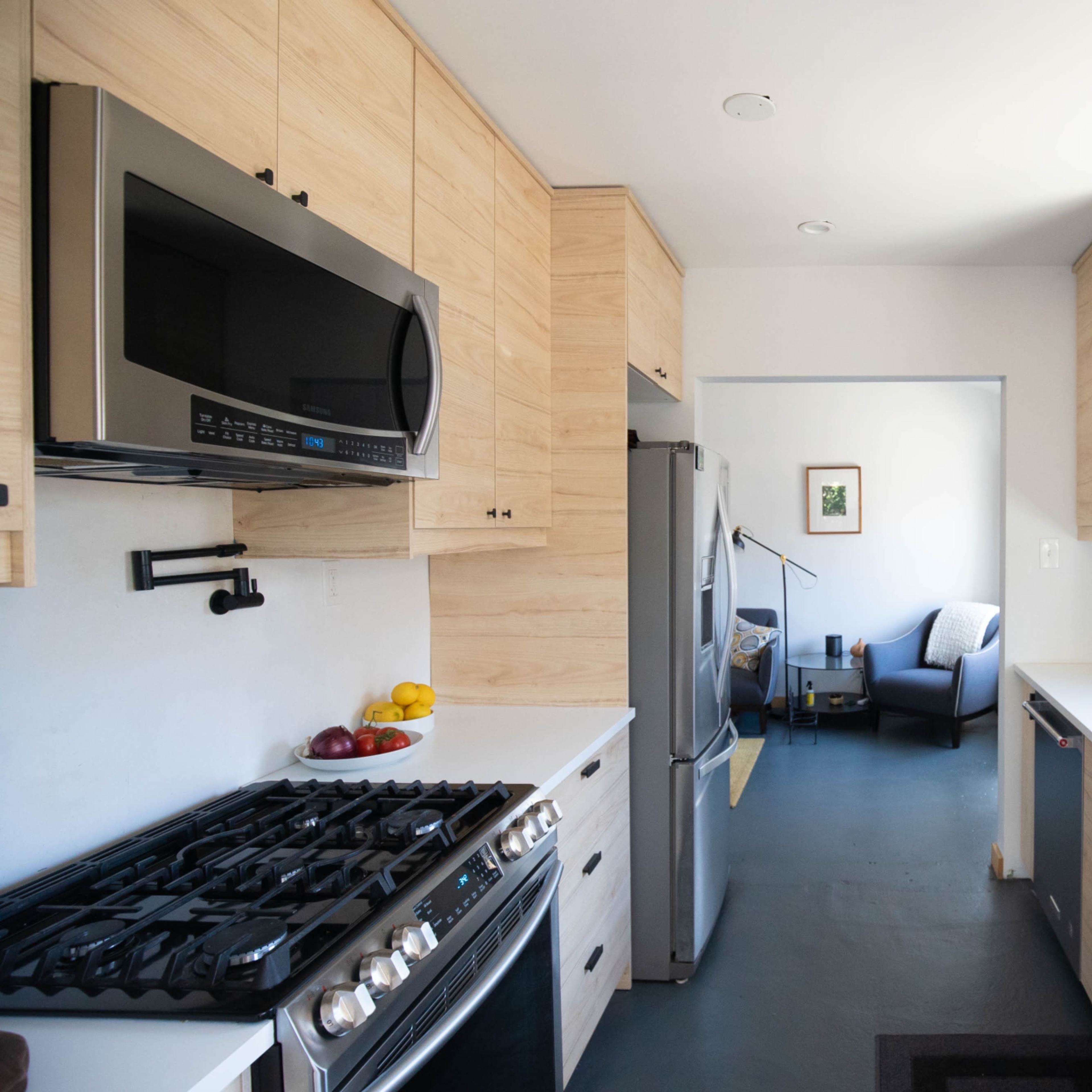 A modern kitchen features wooden cabinetry, stainless steel appliances, and an open layout leading to a living area.