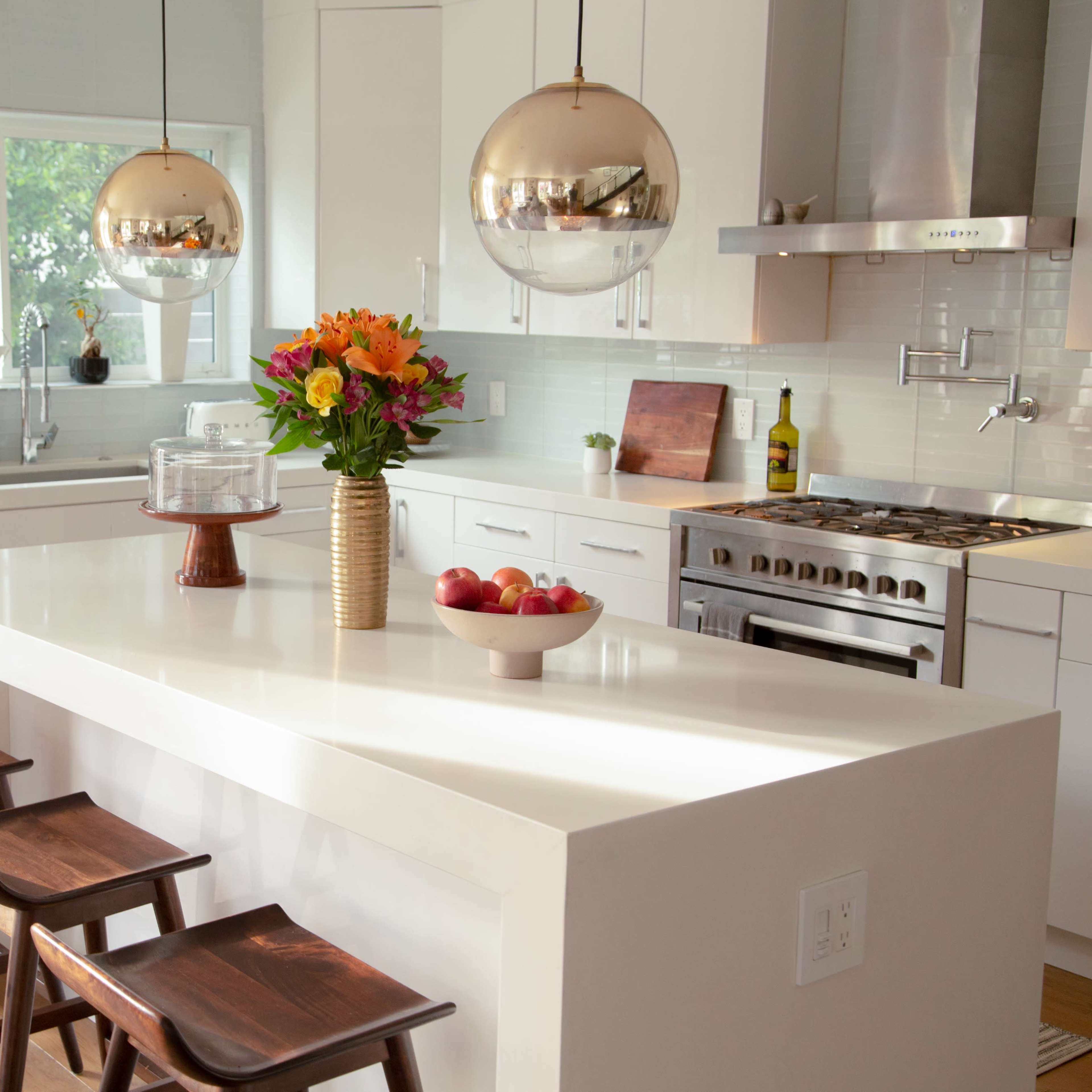 A modern kitchen features white cabinetry, a central island with three wooden stools, and pendant lighting above, adorned with a vase of flowers and a bowl of apples.