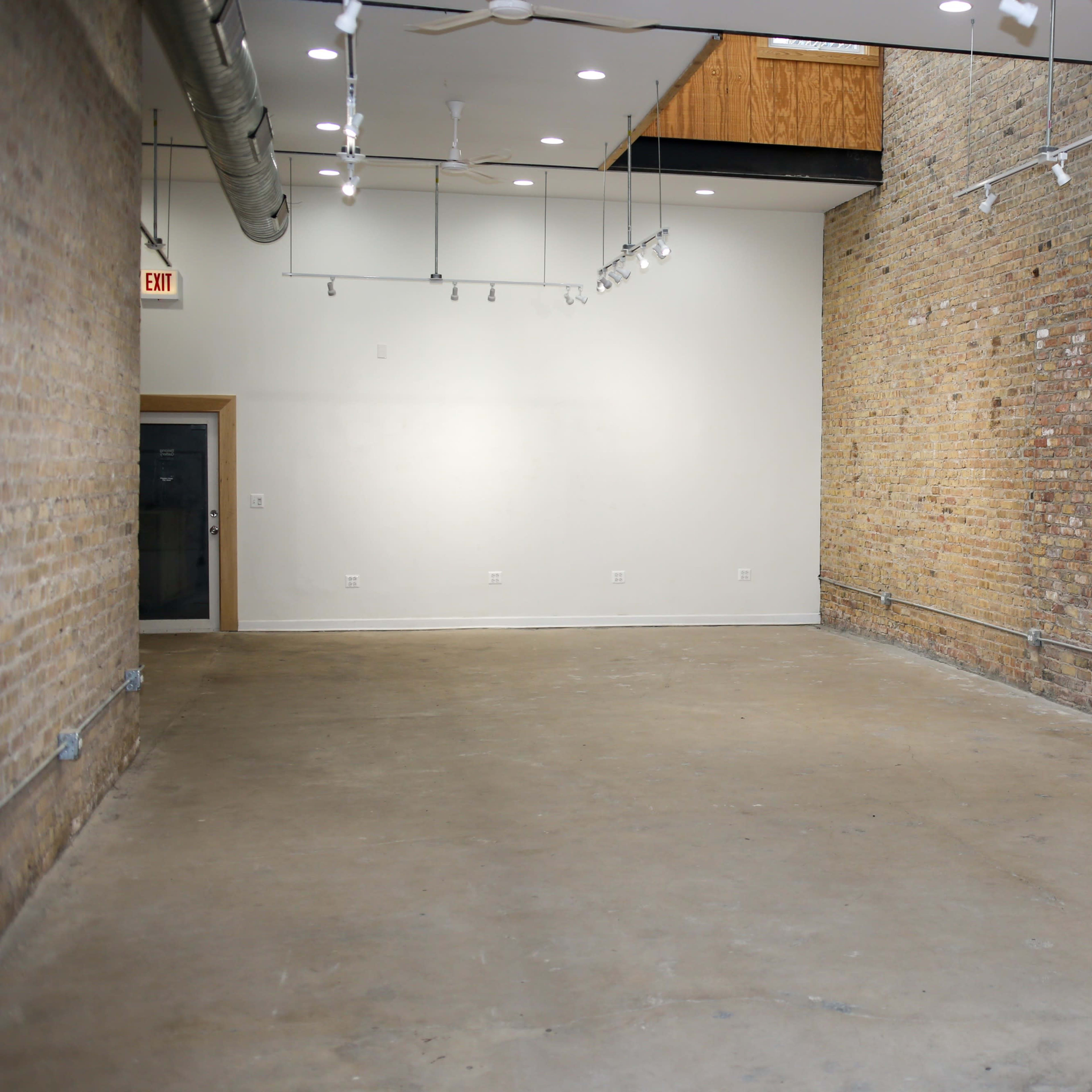 A spacious, empty room with exposed brick walls and concrete flooring, illuminated by track lighting.