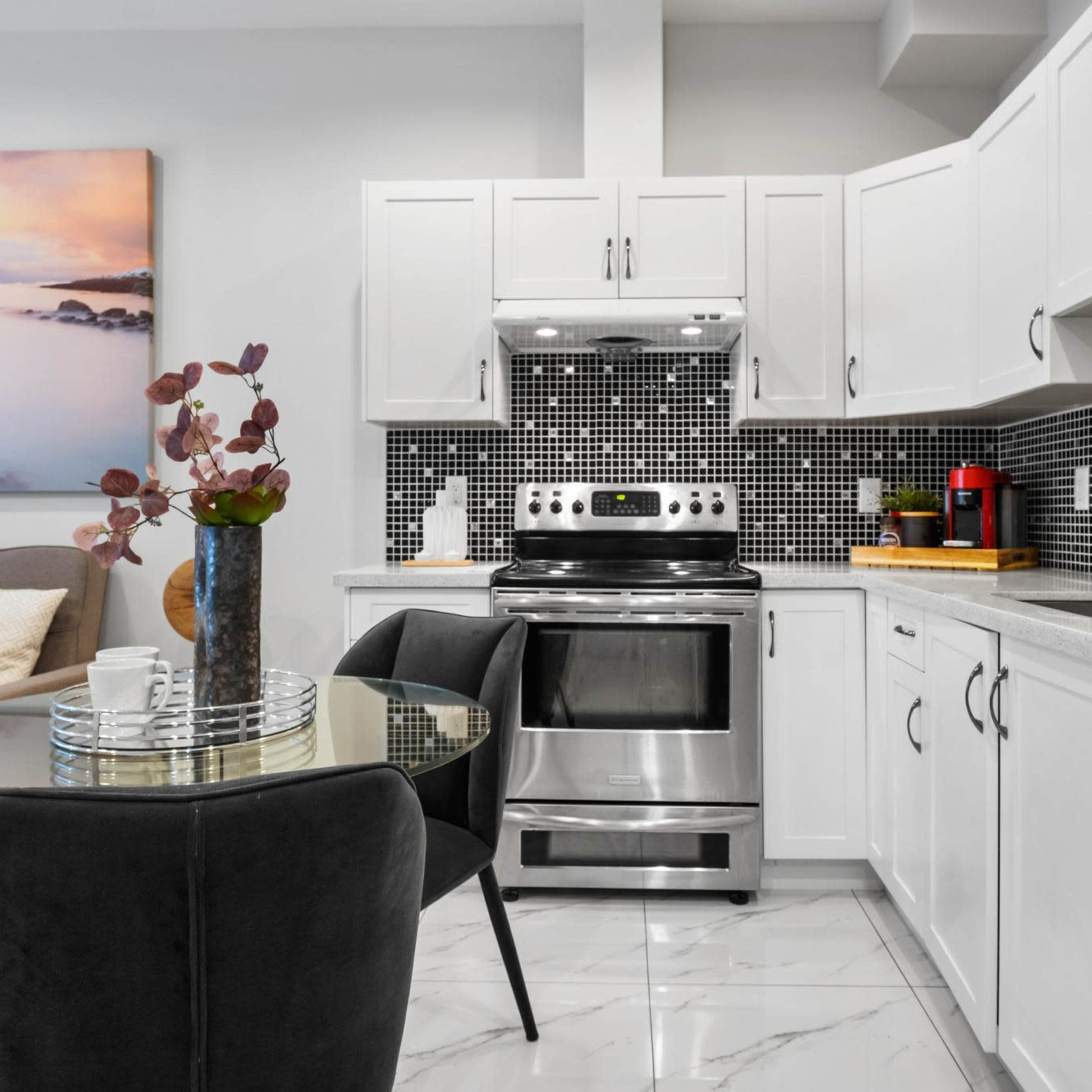 A modern kitchen features white cabinets, a stainless steel stove, and a circular glass dining table with black velvet chairs.