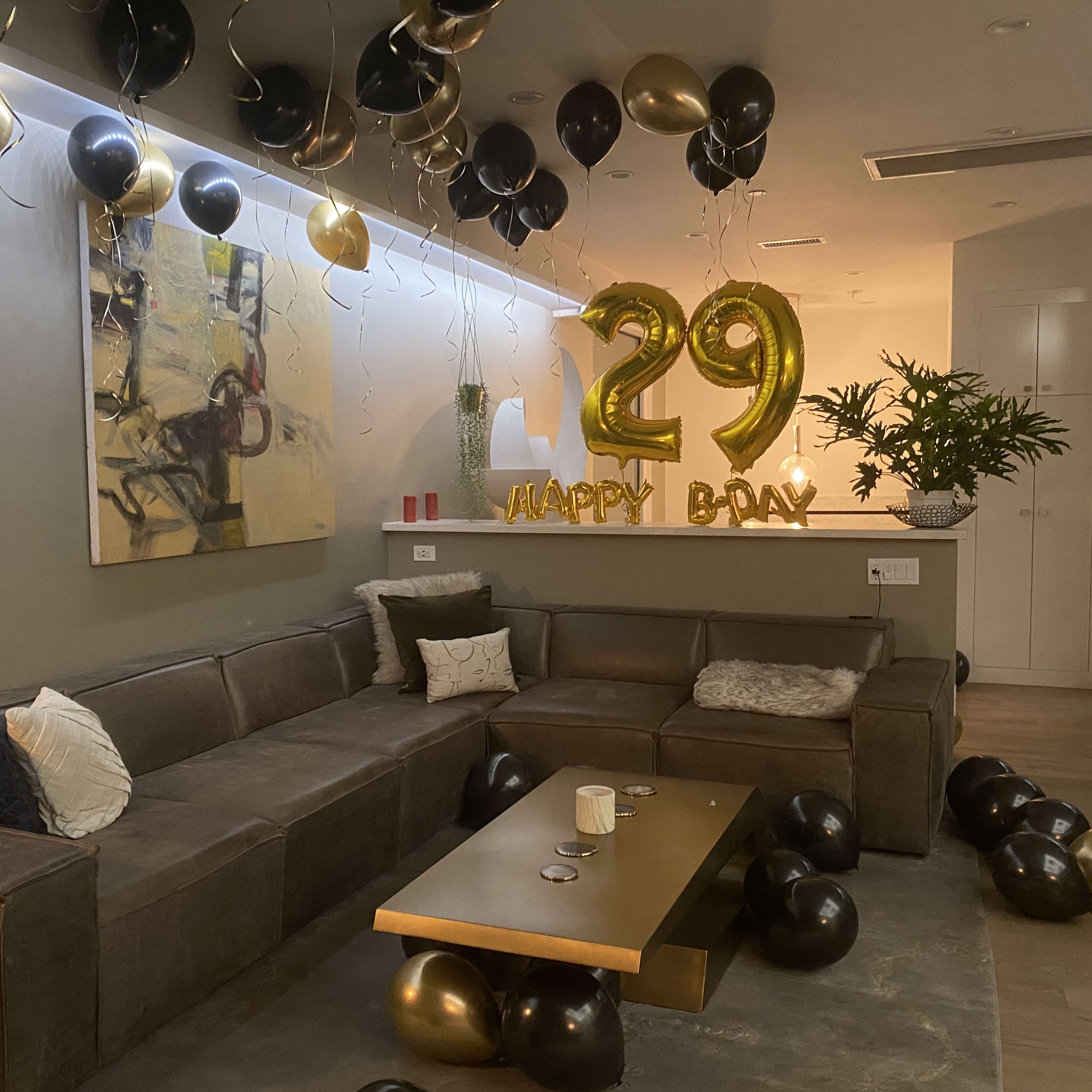 A living room is decorated for a 29th birthday celebration, featuring black and gold balloons along with large gold numeral balloons that spell out "29" and "HAPPY BDAY."