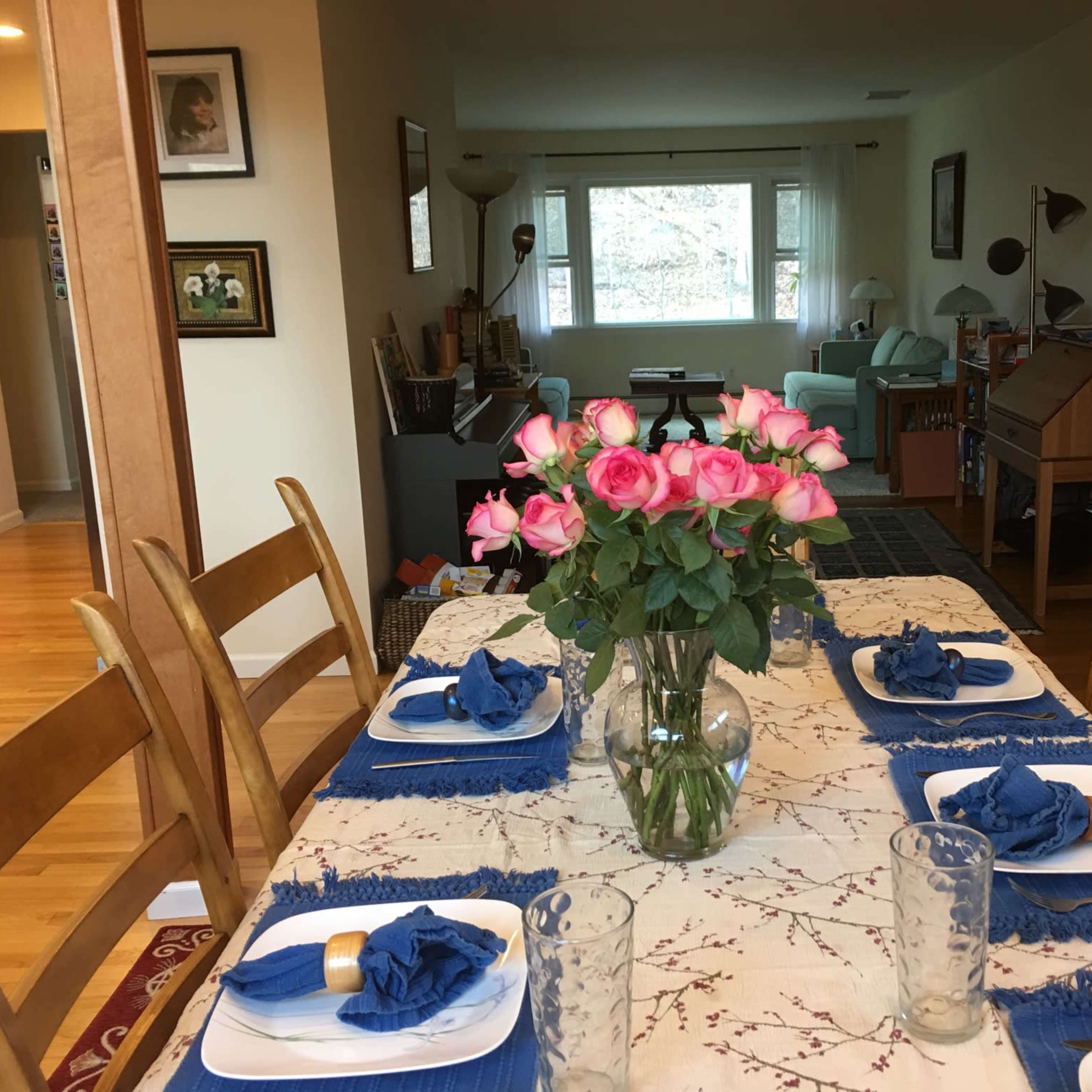 A dining table set with blue napkins and plates is adorned with a vase of pink roses, while a living room with light-colored furniture is visible in the background.