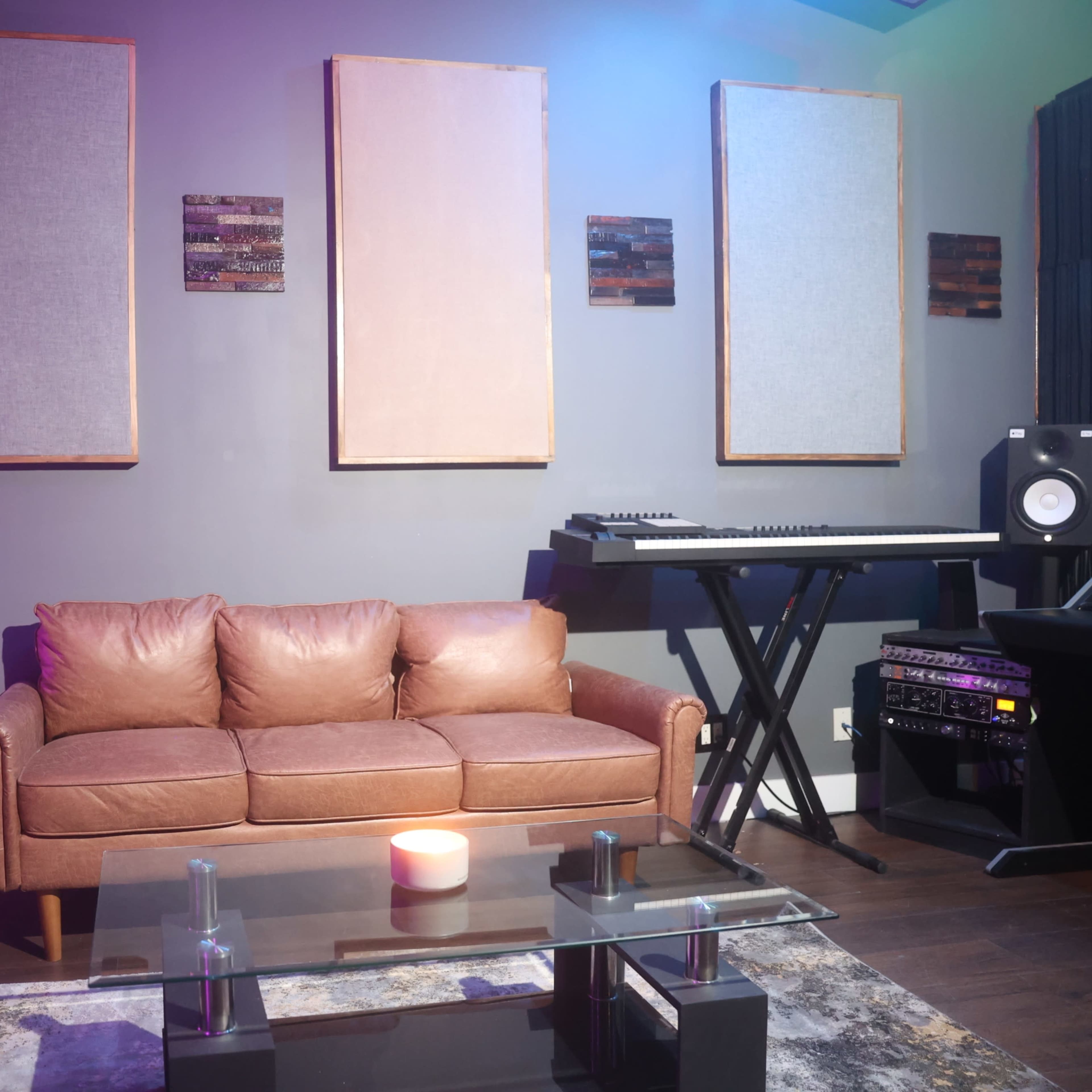 A contemporary lounge area features a leather sofa, a glass coffee table, and a music keyboard setup against a wall with three framed acoustic panels.