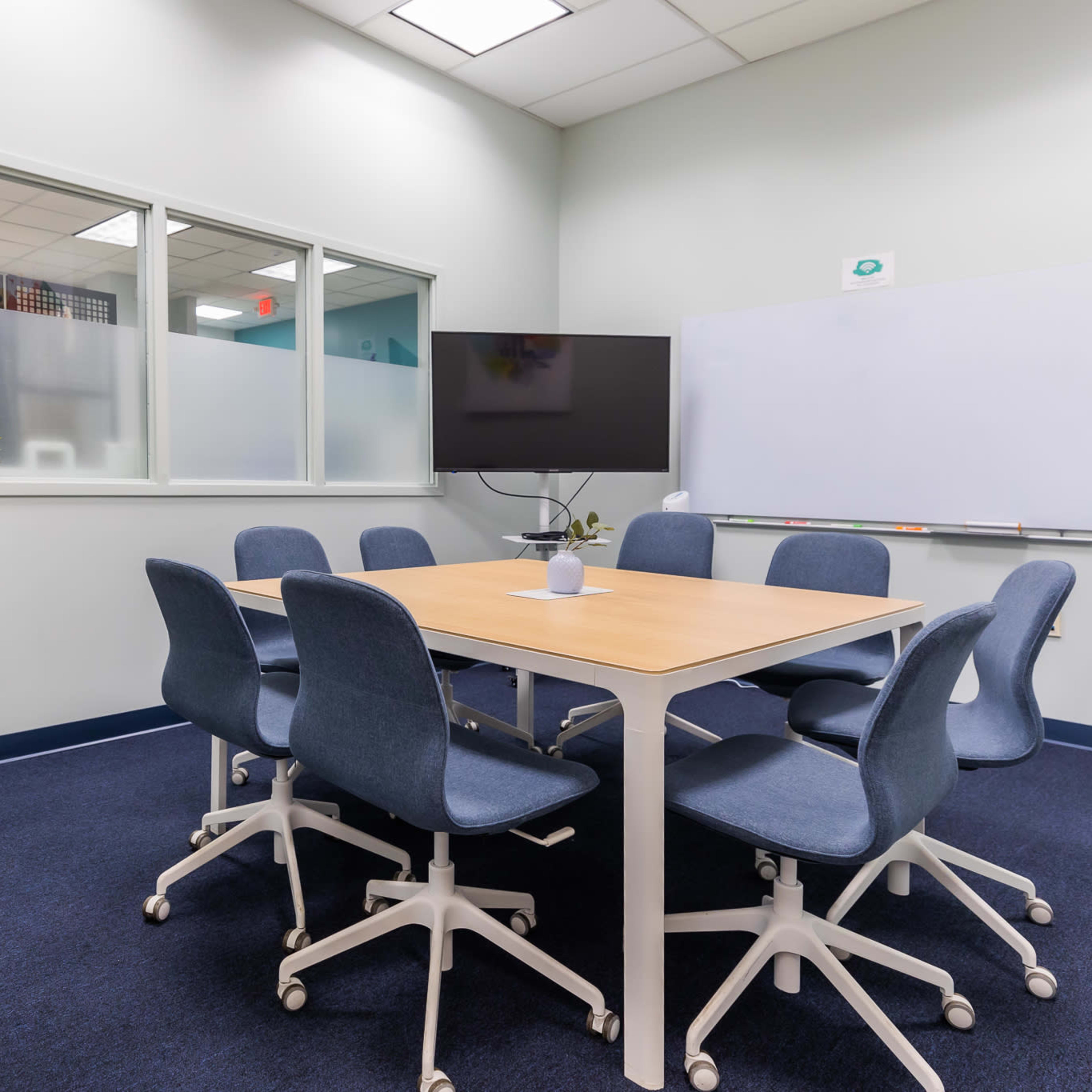 A modern conference room features a wooden table surrounded by eight blue chairs, a wall-mounted screen, a whiteboard, and a potted plant in the corner.