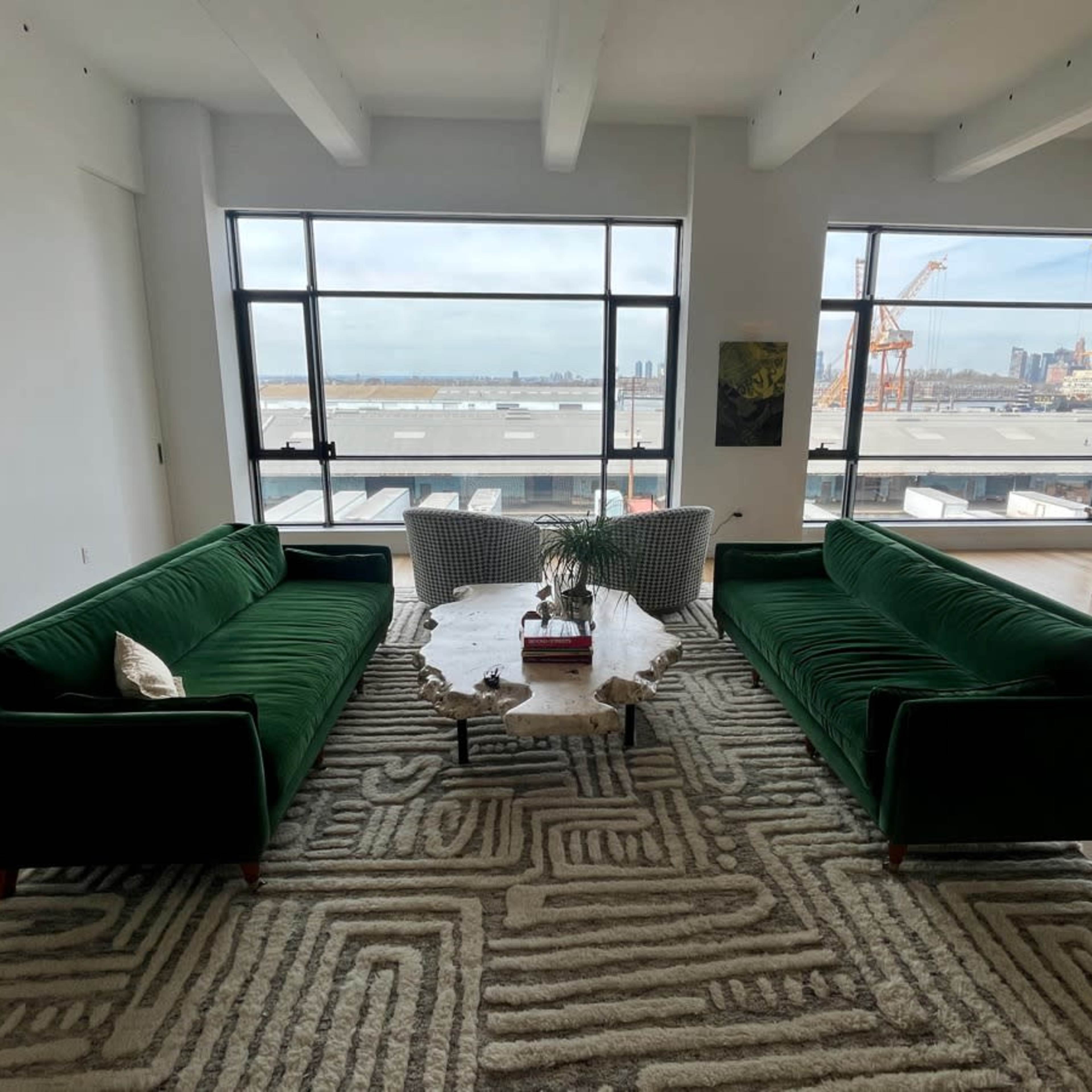 The scene depicts a spacious modern living area with two green velvet sofas facing a coffee table, surrounded by large windows displaying a view of a waterfront and distant city skyline.