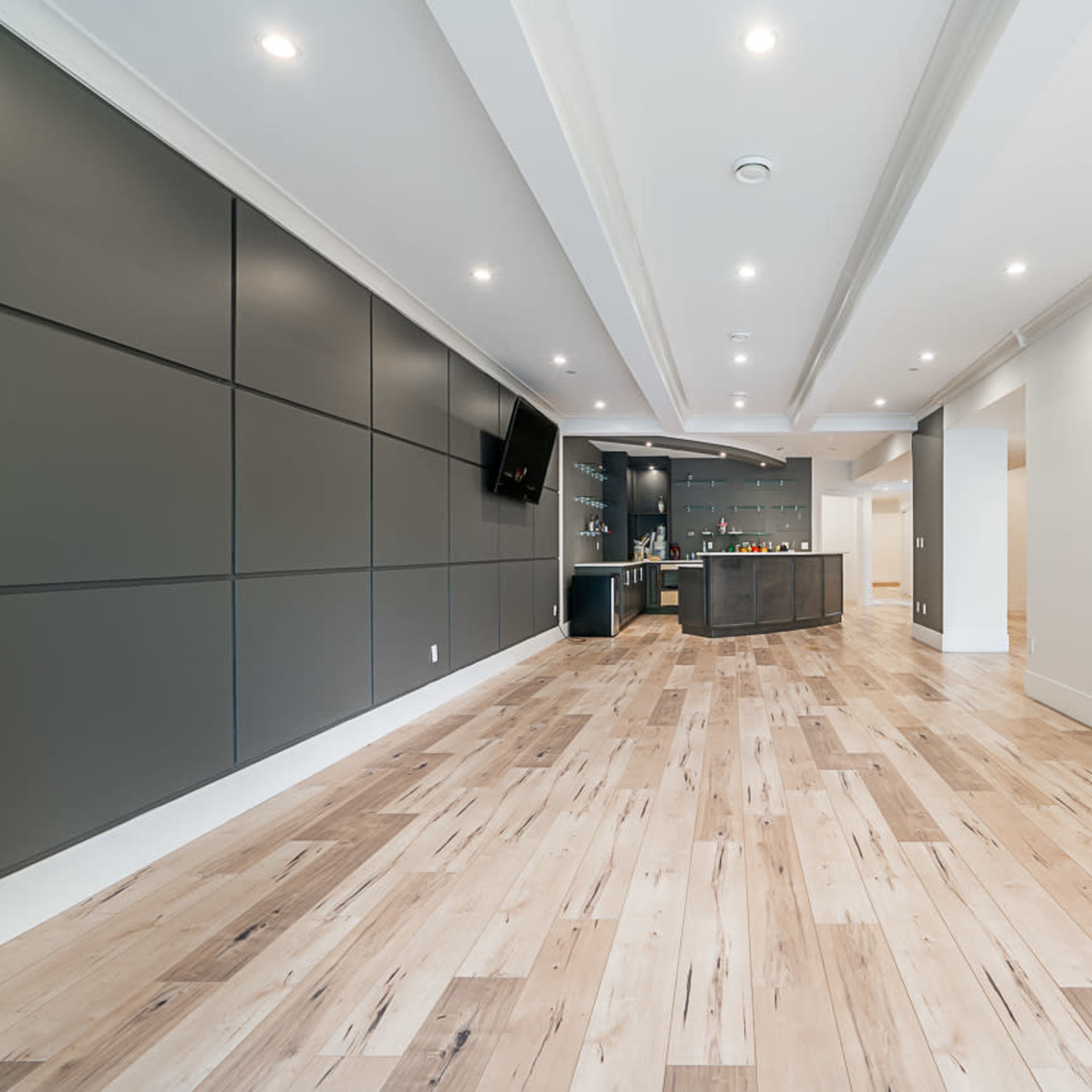 A spacious interior room with wooden flooring, gray panel walls, and a kitchen area featuring a dark countertop and shelves.